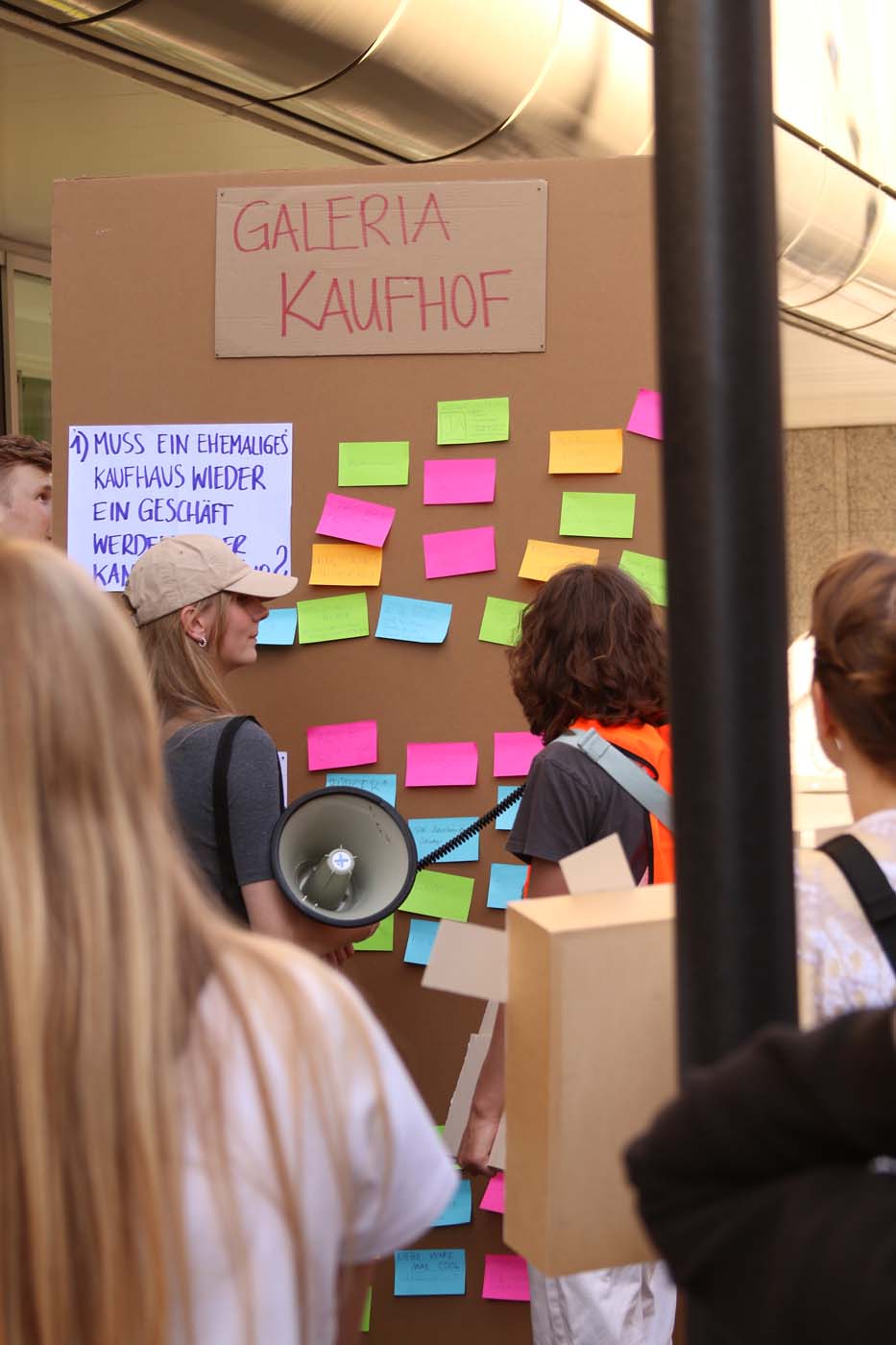 Mehrere Menschen, darunter auch Studierende, stehen vor einer Tafel mit der Aufschrift „Galeria Kaufhof“, die mit bunten Klebezetteln bedeckt ist. Eine Person hält ein Megafon, eine andere einen Pappkarton – die Szene erinnert an einen Stadtspaziergang zum Thema Baukulturverlust. at Hochschule Coburg