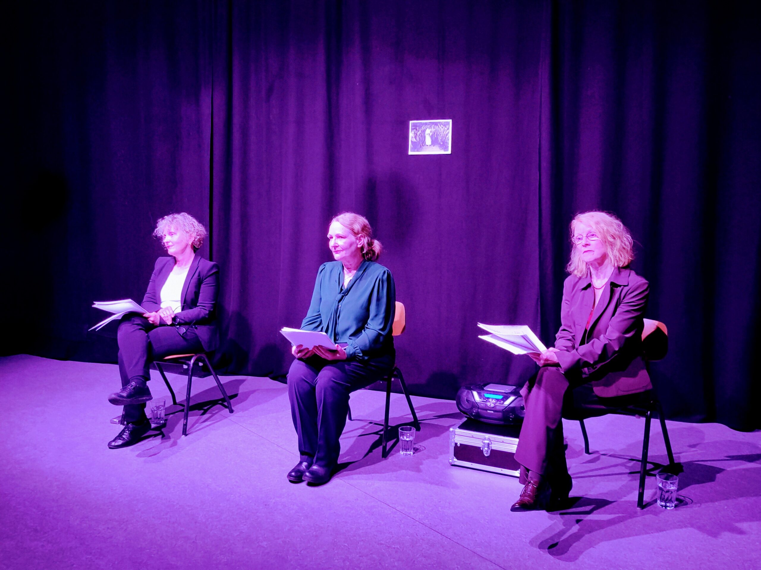 Drei Frauen in Businesskleidung sitzen unter violettem Licht auf der Bühne und halten Unterlagen für eine szenische Lesung in den Händen. Ein kleines Bild ist am schwarzen Vorhang hinter ihnen befestigt und sorgt für eine dramatische Atmosphäre. at Hochschule Coburg