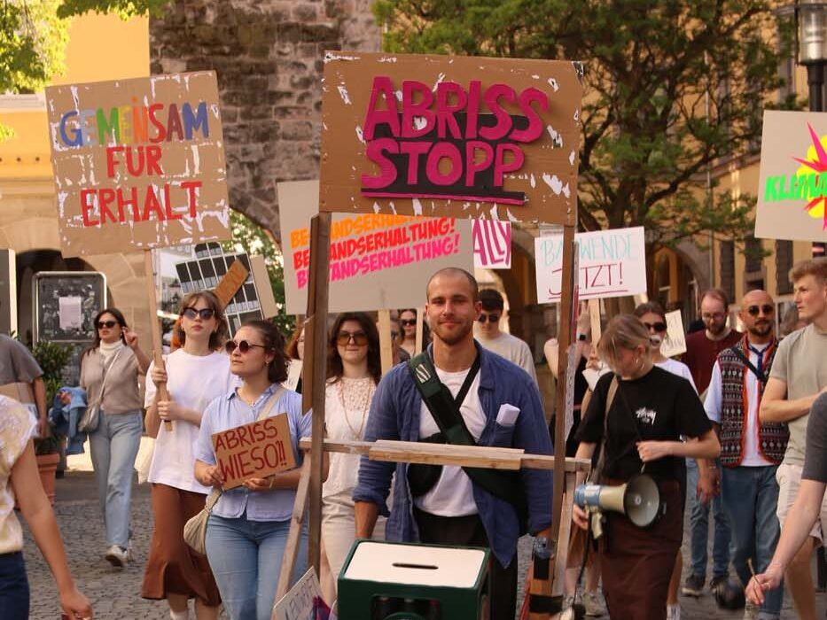Eine Gruppe von Menschen, darunter Studierende, marschiert mit Protestschildern auf einer Kopfsteinpflasterstraße. Ein Mann schiebt eine Schubkarre mit Kisten und einem Koffer. Die Schilder sind mit deutschen Texten versehen und betonen in friedlicher und organisierter Stimmung die Sorge um Baukulturverlust. at Hochschule Coburg
