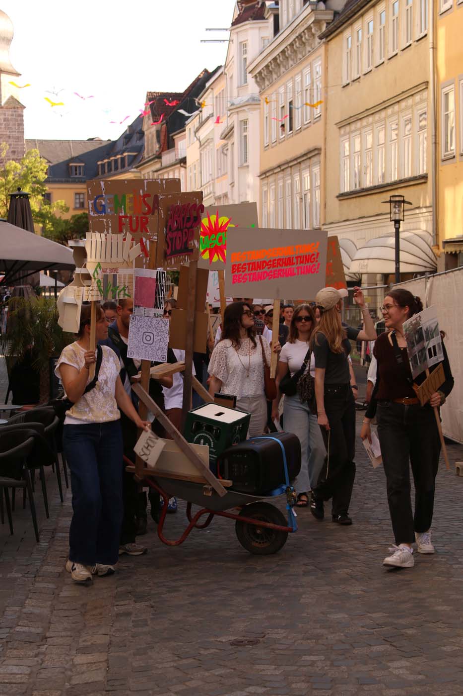 Studierende gehen eine Kopfsteinpflasterstraße entlang und halten bunte Protestschilder – einige auf Deutsch – hoch, die auf Probleme wie Baukulturverlust hinweisen. Eine Person schiebt eine Schubkarre mit einem Lautsprecher, während sie ihren Stadtspaziergang an Gebäuden und Sitzgelegenheiten im Freien vorbei fortsetzen. at Hochschule Coburg
