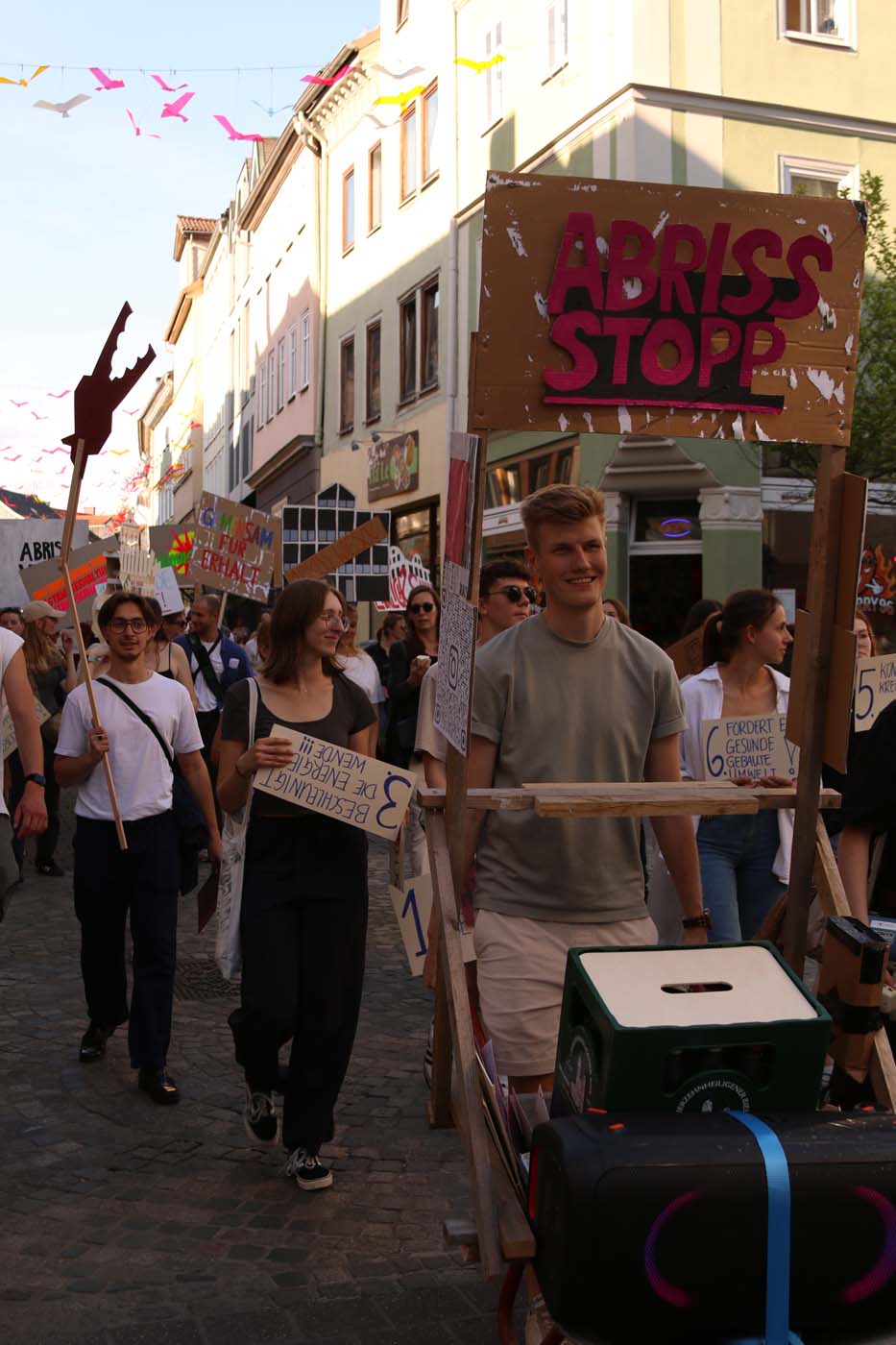 Eine Gruppe Studierender marschiert mit Protestschildern eine Straße entlang. Auf einem großen Schild steht „ABRISS STOPP“ und macht auf den Verlust von Baukultur aufmerksam. Die Atmosphäre wirkt friedlich und energiegeladen. at Hochschule Coburg