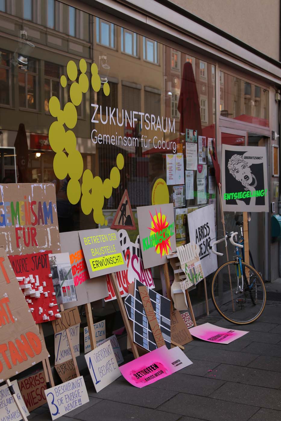 Ein Schaufenster mit dem Schild „Zukunftsraum Gemeinsam für Coburg“ zeigt bunte Protestschilder und Plakate zum Thema Baukulturverlust. In der Nähe ruht ein Fahrrad, während Stadtspaziergang-Reflexionen und Studierende Leben in die Szene bringen. at Hochschule Coburg