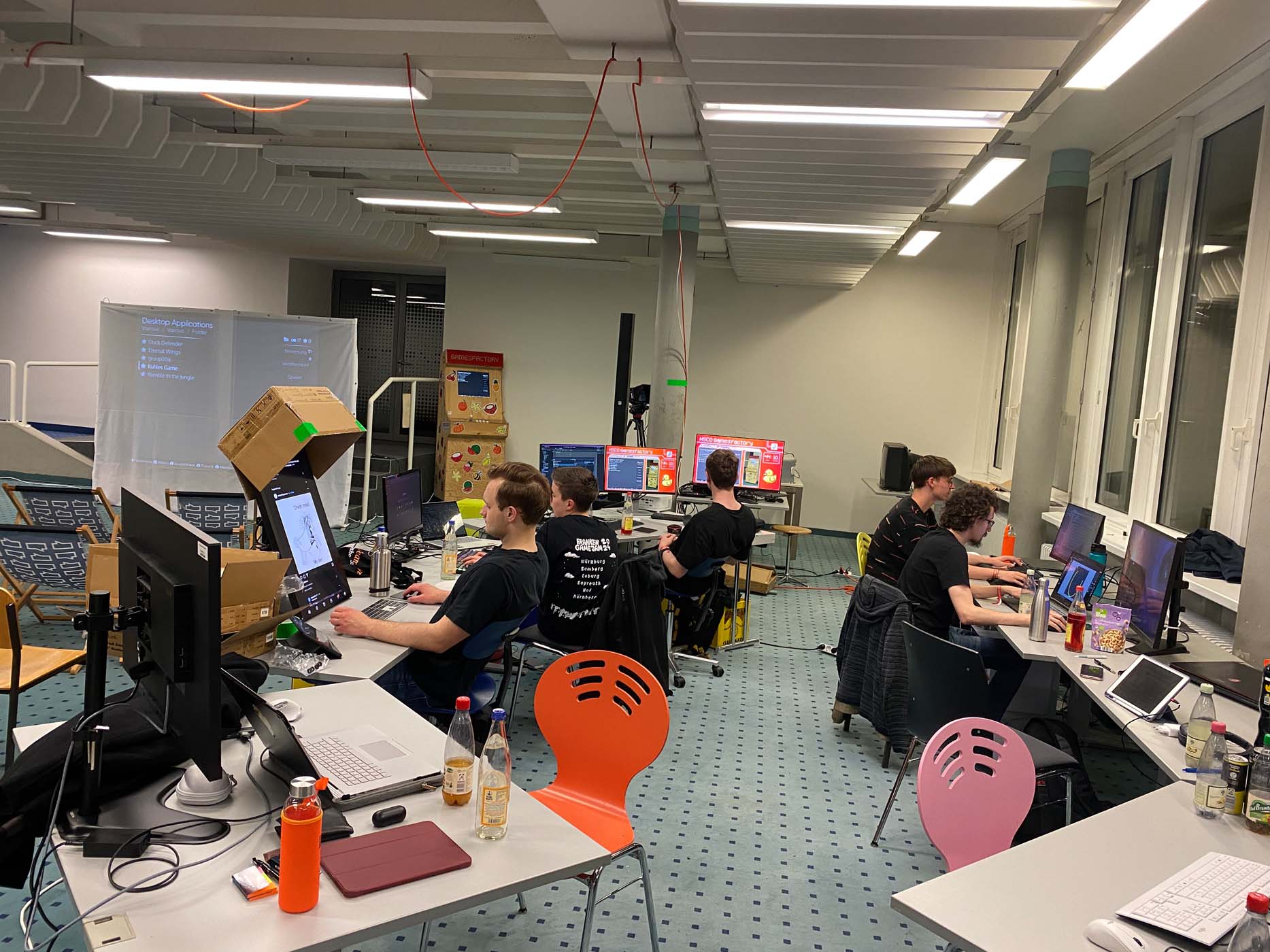 Eine Gruppe von Menschen arbeitet während eines Game Jams an Computern in einem hell erleuchteten, modernen Büro. Die Schreibtische sind voller Bildschirme, Getränke und Kabel; einige Monitore sind von Pappkartons umgeben. Die Atmosphäre ist geschäftig und konzentriert. at Hochschule Coburg