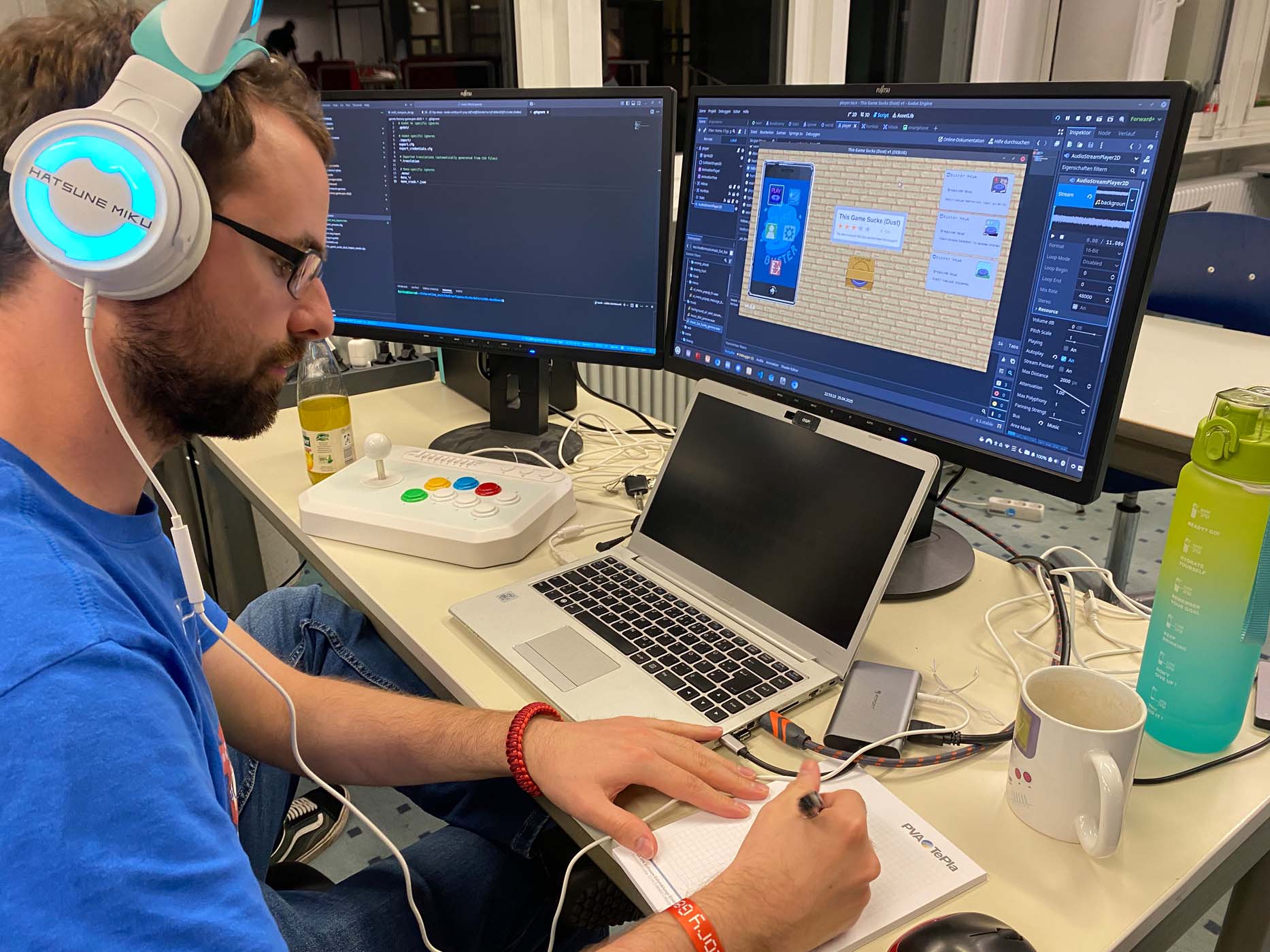 Eine Person mit leichtem Gepäck und Kopfhörern arbeitet an einem Schreibtisch mit zwei Monitoren, einem Laptop, einem Gamecontroller, einer Wasserflasche und einer Kaffeetasse und macht sich Notizen. Ein Bildschirm zeigt Code, der andere eine Game-Jam-Designoberfläche. at Hochschule Coburg