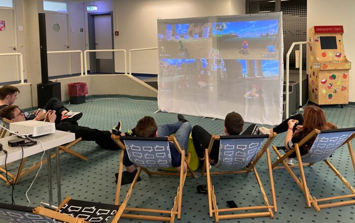 Fünf Personen mit leichtem Gepäck entspannen auf Liegestühlen in einem Innenraum und schauen und spielen ein Videospiel, das auf eine große weiße Leinwand projiziert wird. Rechts daneben steht ein Spielautomaten. Die Atmosphäre ist locker und verspielt. at Hochschule Coburg