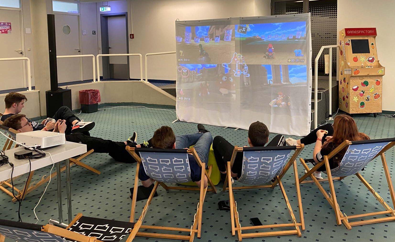 Fünf Personen mit leichtem Gepäck entspannen auf Liegestühlen in einem Innenraum und schauen und spielen ein Videospiel, das auf eine große weiße Leinwand projiziert wird. Rechts daneben steht ein Spielautomaten. Die Atmosphäre ist locker und verspielt. at Hochschule Coburg