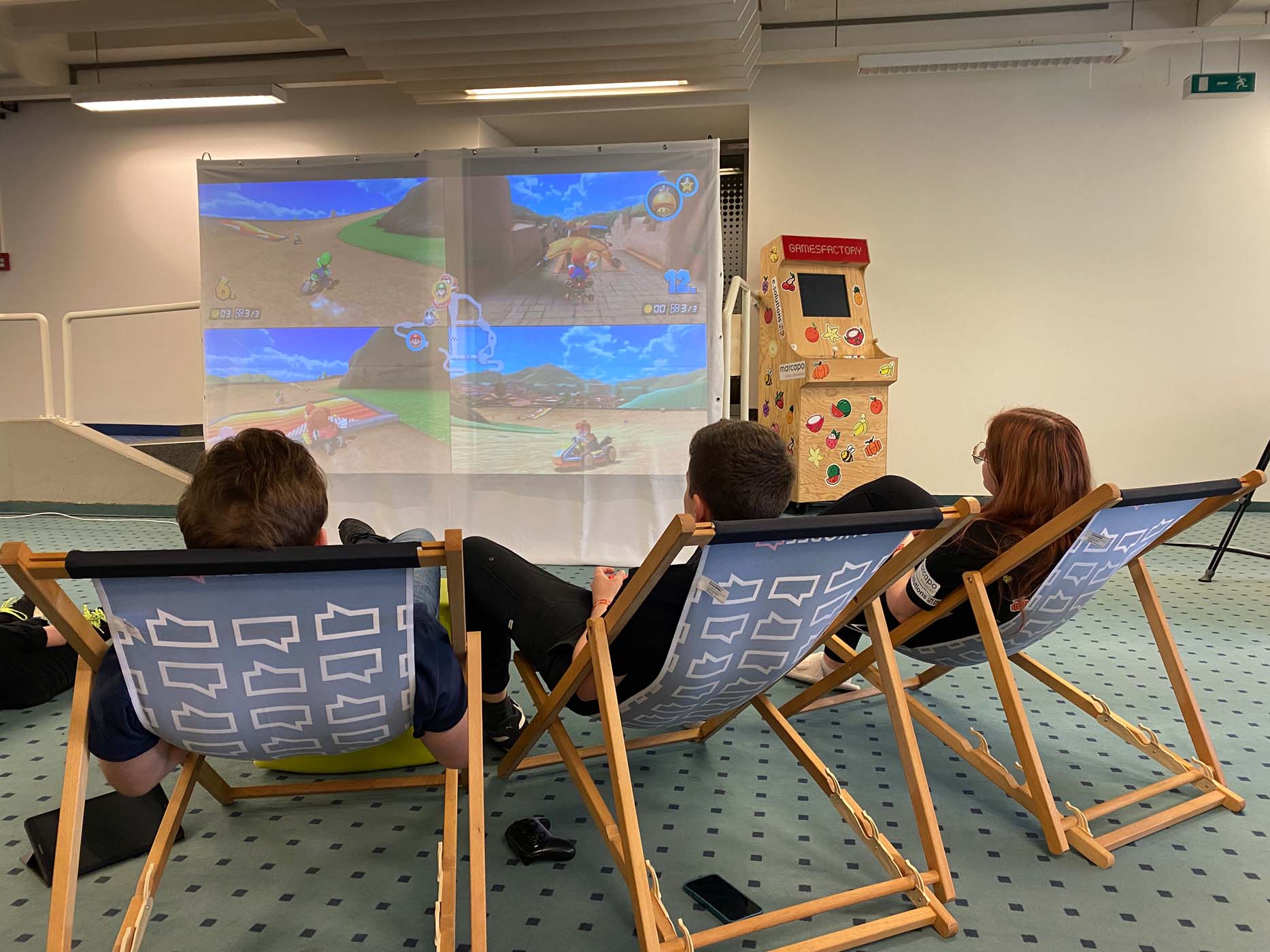Drei Personen sitzen auf Liegestühlen und genießen einen Game Jam, während auf einer großen Leinwand Rennen laufen. Im Hintergrund leuchtet ein Arcade-Automat und sorgt für eine entspannte Lichtreise-Atmosphäre – perfekt für alle, die mit leichtem Gepäck reisen. at Hochschule Coburg