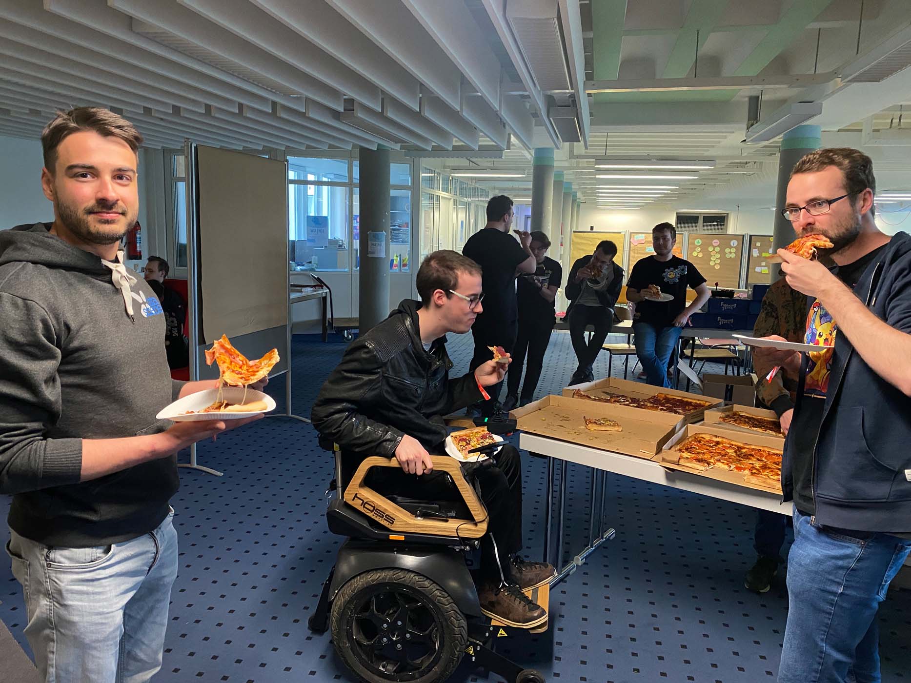 Eine Gruppe von Menschen genießt während eines Game Jams Pizza in einer Büroumgebung. Eine Person im Rollstuhl hält einen Teller, während andere stehen und essen. Offene Pizzakartons stehen auf dem Tisch und schaffen so eine entspannte und freundliche Atmosphäre. at Hochschule Coburg