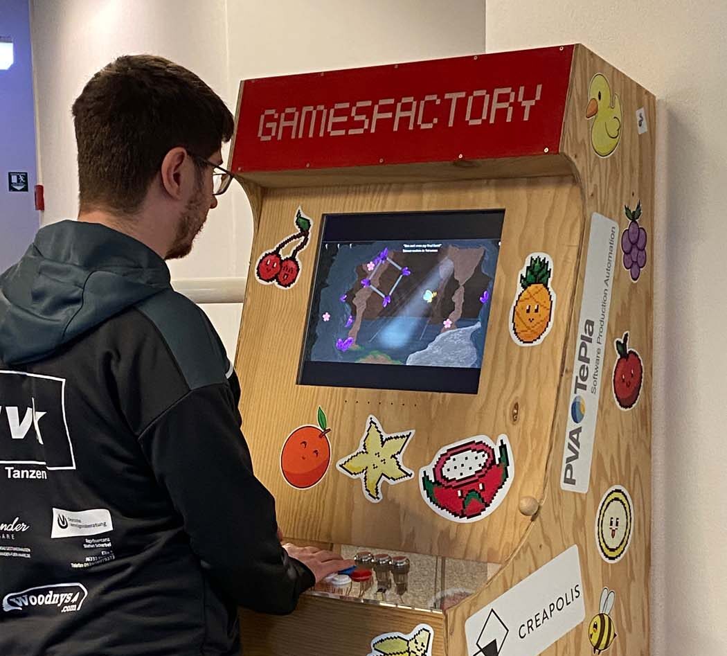 Eine Person mit leichtem Gepäck spielt ein Videospiel an einem hölzernen Arcade-Automaten mit der Aufschrift „GAMESFACTORY“, der mit bunten Fruchtaufklebern und verschiedenen Sponsorenlogos, darunter Creapolis und PVA Tepla, verziert ist. at Hochschule Coburg