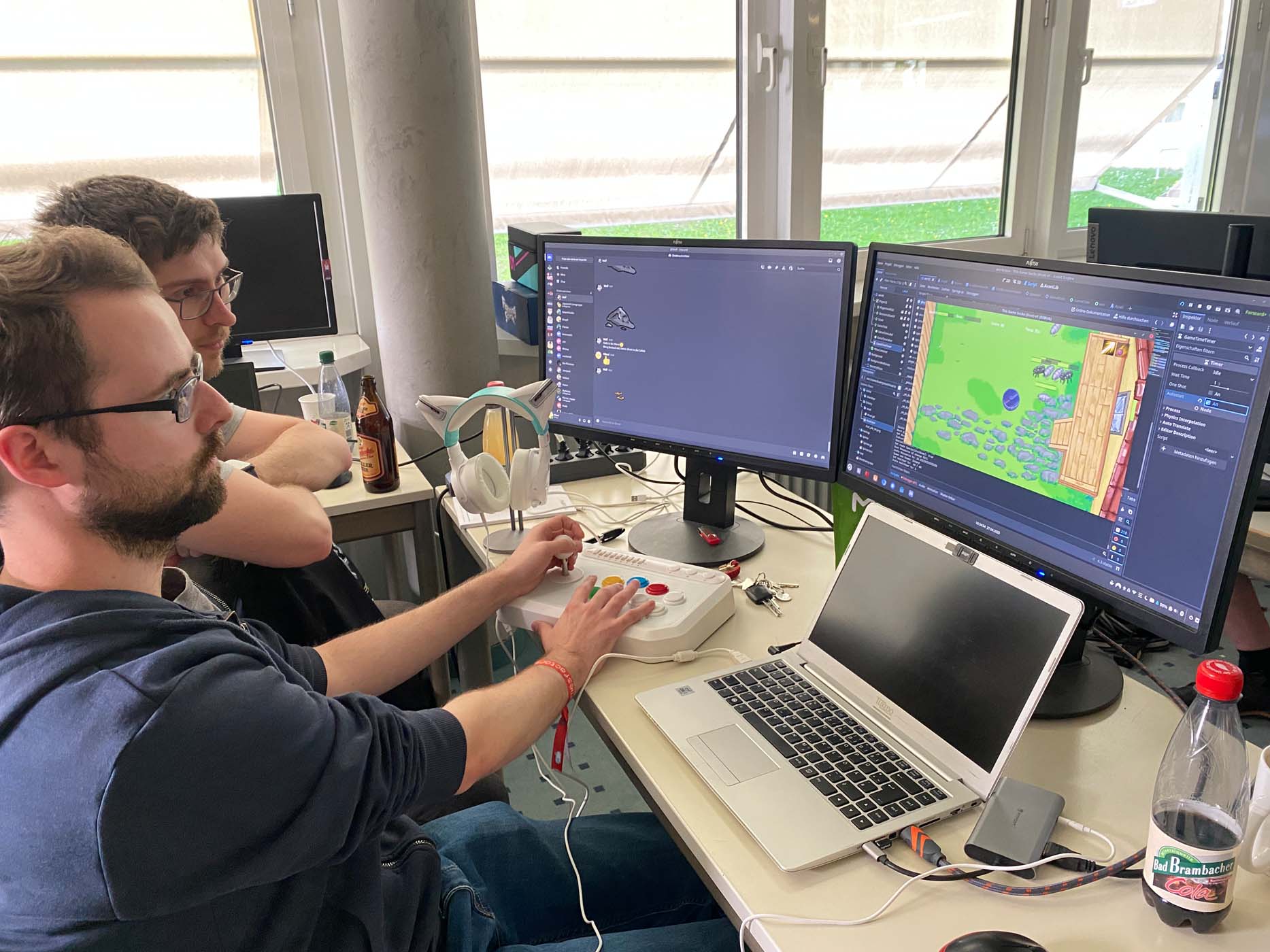 Zwei Personen sitzen an einem Schreibtisch mit mehreren Monitoren, einem Laptop und verschiedenen Getränken. Ganz im Stil des Game Jam bearbeitet einer mit einem Arcade-Controller eine bunte Spielszene, während der andere aufmerksam zusieht – bereit für kreative Lichtreisen mit leichtem Gepäck. at Hochschule Coburg