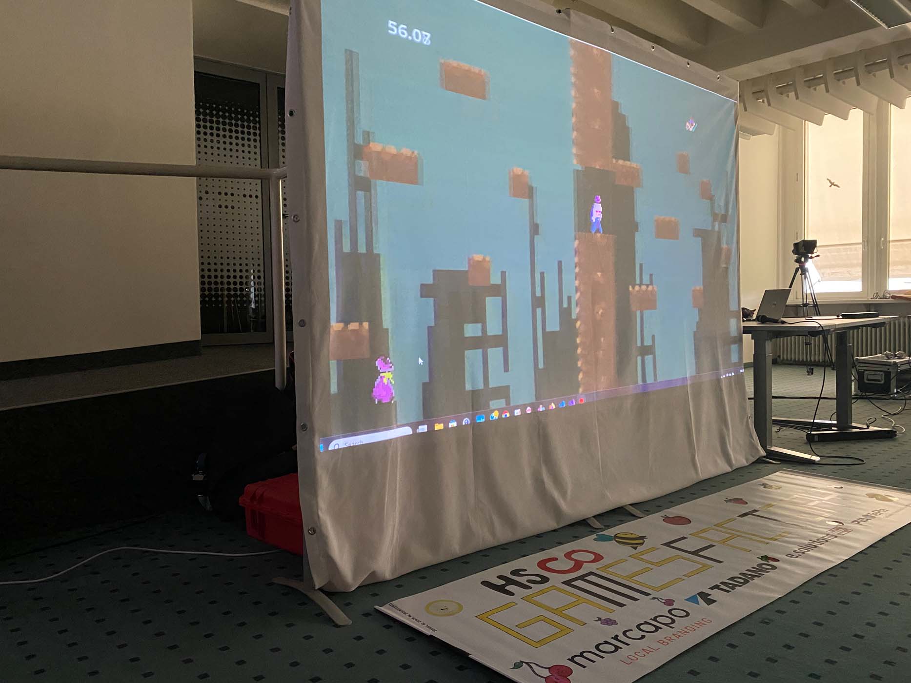 In einem Raum wird ein Videospiel auf eine Leinwand projiziert, auf der verpixelte Figuren auf Plattformen zu sehen sind. Unter der Leinwand ist ein Banner für das „HSCO GAME FEST“ zu sehen, auf dem die Logos der Sponsoren – darunter Lichtreisen – und eine Kamera auf einem Stativ zu sehen sind. at Hochschule Coburg