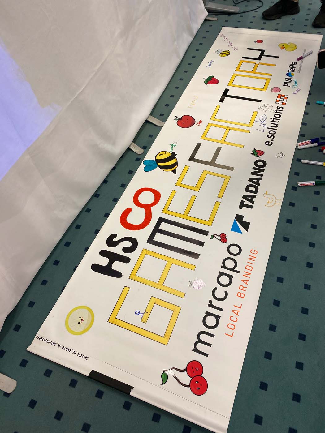 Ein großes weißes Banner auf blauem Teppich trägt die Aufschrift „HS co GAMES FACTORY“ in bunten Buchstaben, Zeichnungen einer Biene, Würfeln und Spielmarken – und fängt damit den Geist des Game Jam ein. Sponsorenlogos wie Marcapo und TADANO sind zu sehen, und daneben sind farbige Markierungen verteilt. at Hochschule Coburg