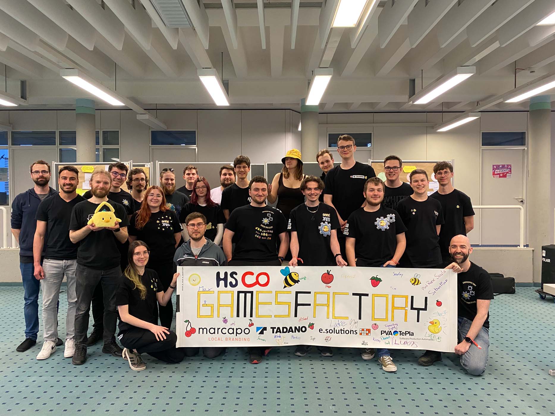 Eine Gruppe von etwa 22 Personen posiert lächelnd im Innenbereich und hält ein großes, buntes „HS CO GamesFactory“-Banner mit Sponsorenlogos und verspielten Illustrationen hoch. Die meisten tragen schwarze Shirts und feiern gemeinsam ihren kreativen Game-Jam-Geist. at Hochschule Coburg