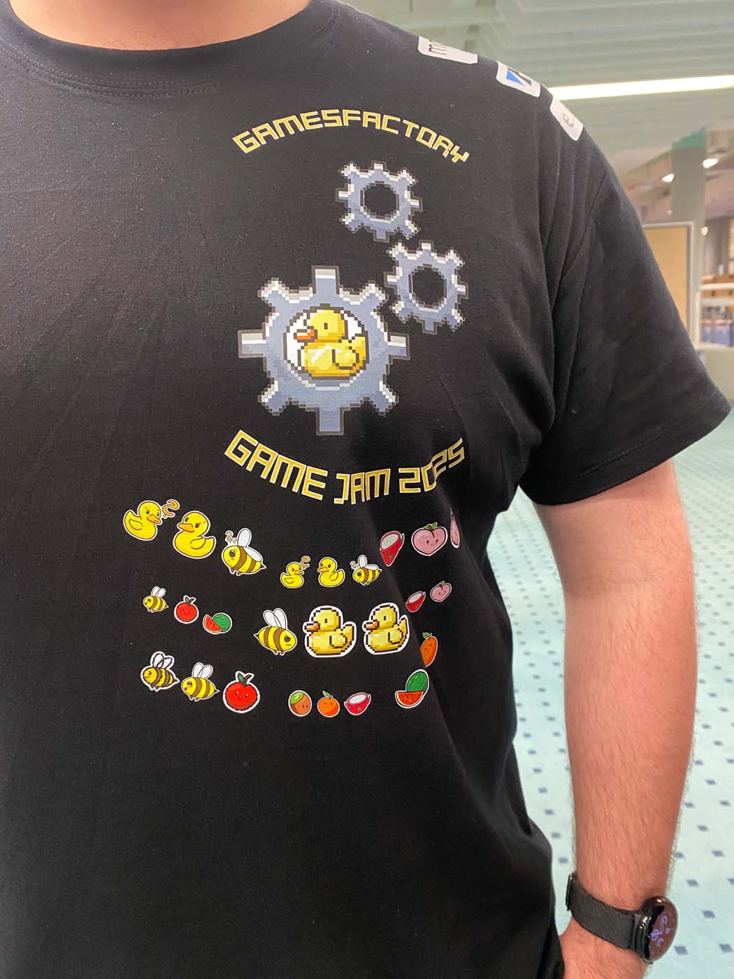 Eine Person, die ein schwarzes T-Shirt mit Pixel-Art-Zahnrädern, Enten und Früchten und der gelben Aufschrift „GAMESFACTORY GAME JAM 2025“ trägt, steht in einem öffentlichen Innenraum mit blauem Teppich – bereit für den Game Jam mit leichtem Gepäck und kreativem Geist. at Hochschule Coburg