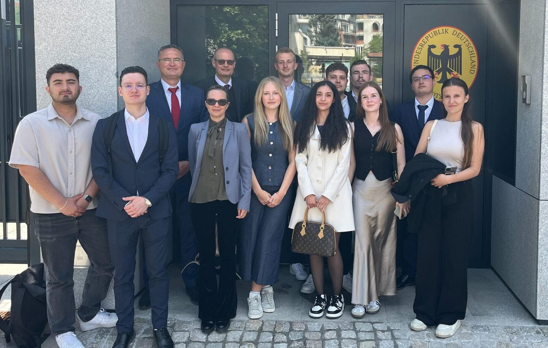 Eine Gruppe von dreizehn Personen in Business- und Freizeitkleidung steht gemeinsam vor einem modernen Gebäude mit Steinmauern und einem Schild mit der Aufschrift „Deutsch-Bulgarisches Studien-Projekt“ vor der Tür des deutschen Konsulats bzw. der Botschaft. at Hochschule Coburg