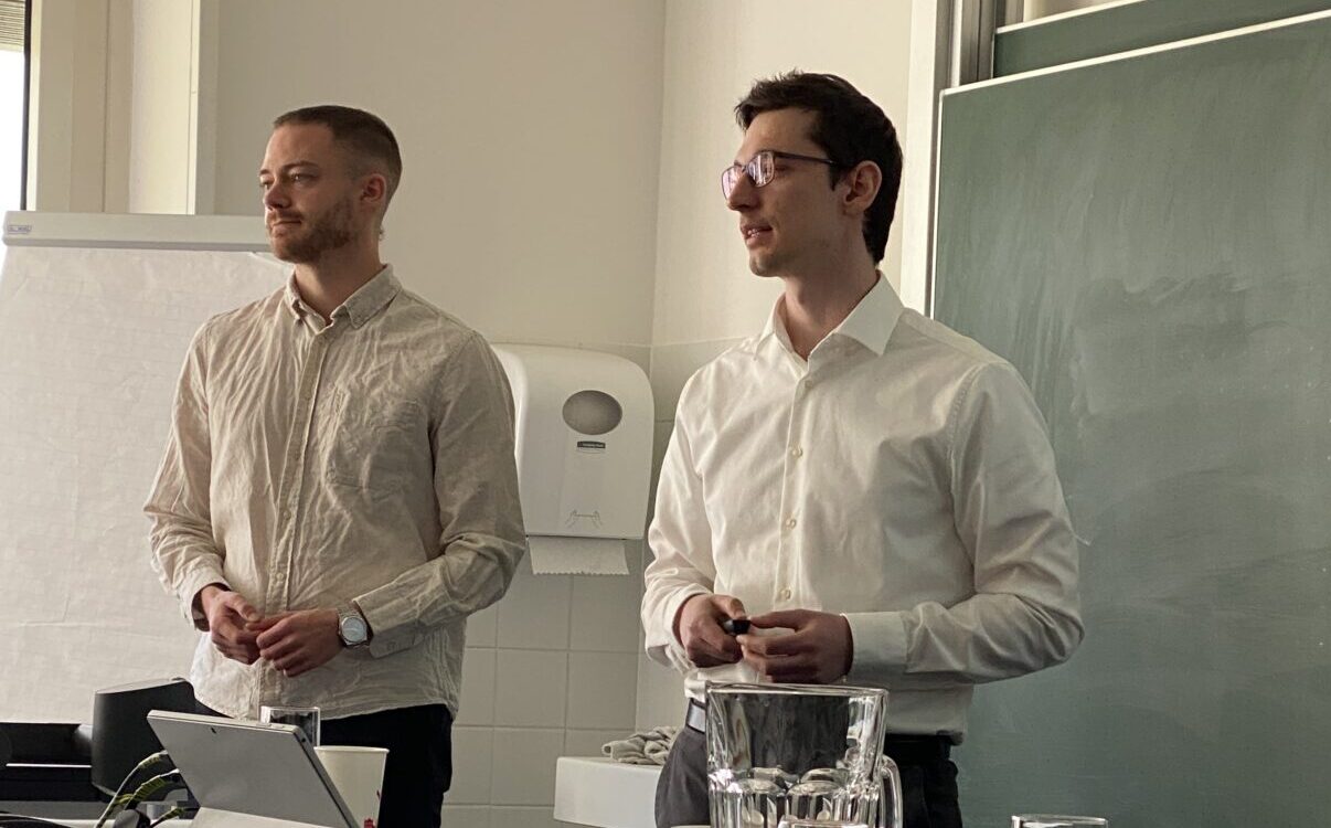 Zwei Männer in weißen Hemden stehen vor einem Klassenraum neben einer Tafel und einem Flipchart und diskutieren über Grüne Geldanlagen. Auf dem Tisch stehen ein Wasserkrug, Gläser, ein Laptop und ein buntes Tuch. Der Raum wirkt hell und professionell. at Hochschule Coburg