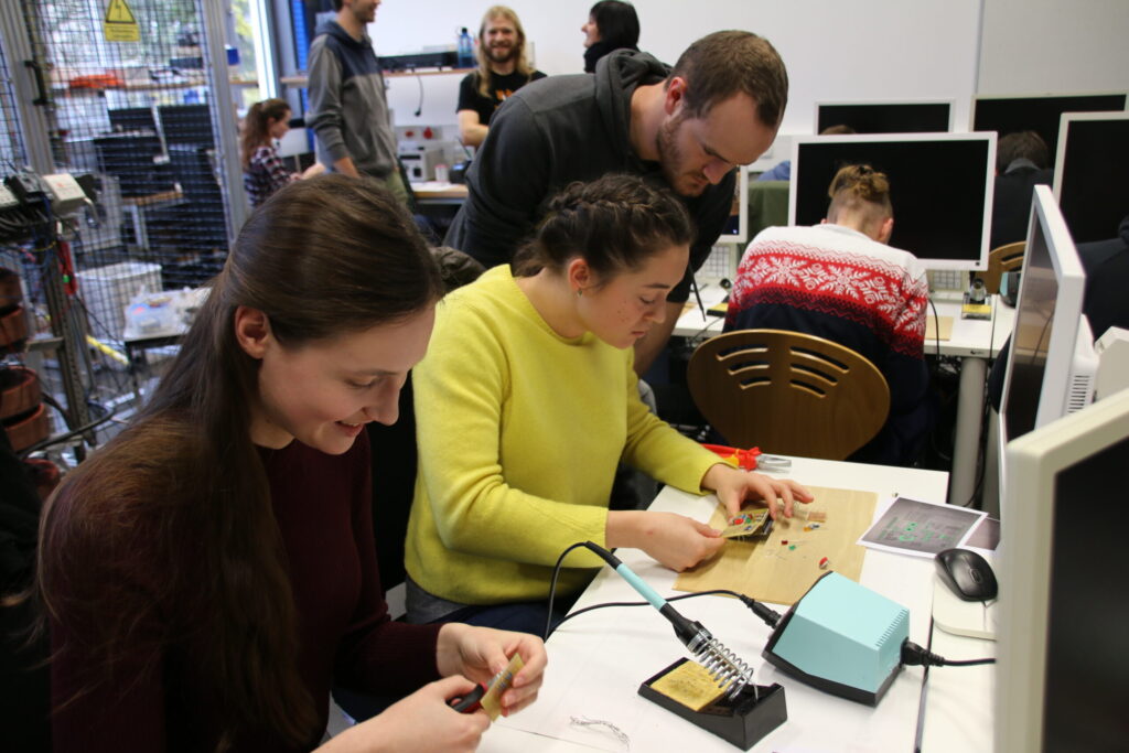 Three people sit at a table working on electronics, using soldering irons and parts. Others stand and talk in the background. Computer monitors, tools, and equipment are visible throughout the busy classroom or workshop. at Hochschule Coburg