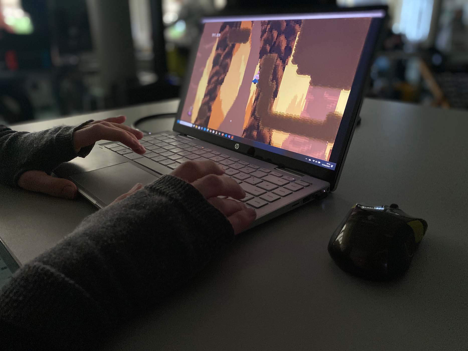 Eine Person in einem dunklen Langarmshirt spielt auf einem silbernen HP-Laptop ein Videospiel im Pixel-Art-Stil, das während eines Game Jams entwickelt wurde. Eine schwarze Computermaus liegt neben dem Laptop auf dem Schreibtisch. at Hochschule Coburg