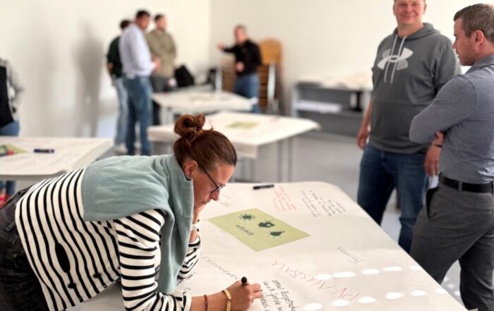 Eine Frau schreibt auf einem großen Blatt Papier auf einem Tisch, während mehrere Personen in einem hellen, modernen Hörsaal der Hochschule Coburg stehen und sich unterhalten. Die kollaborative Atmosphäre spiegelt ihren gemeinsamen Fokus auf Klimaneutralität bis 2040 wider. at Hochschule Coburg
