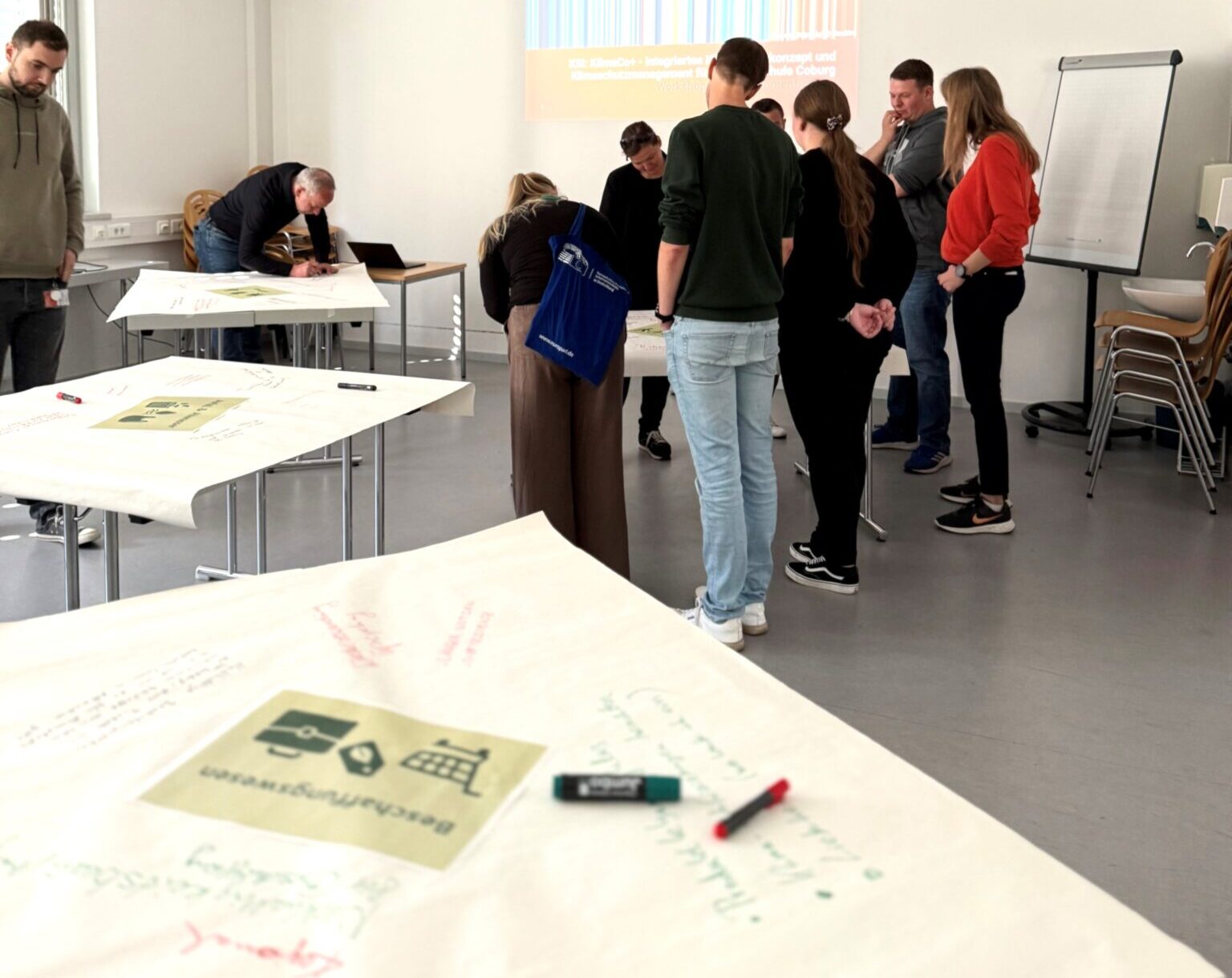 Eine Gruppe von Menschen steht in einem hellen Hörsaal der Hochschule Coburg an einem Tisch und diskutiert. Eine Präsentation zum Thema Klimaneutralität 2040 wird an die Wand projiziert, während Notizen und Diagramme auf Papier sichtbar sind, während die Teilnehmer schreiben oder lesen. at Hochschule Coburg