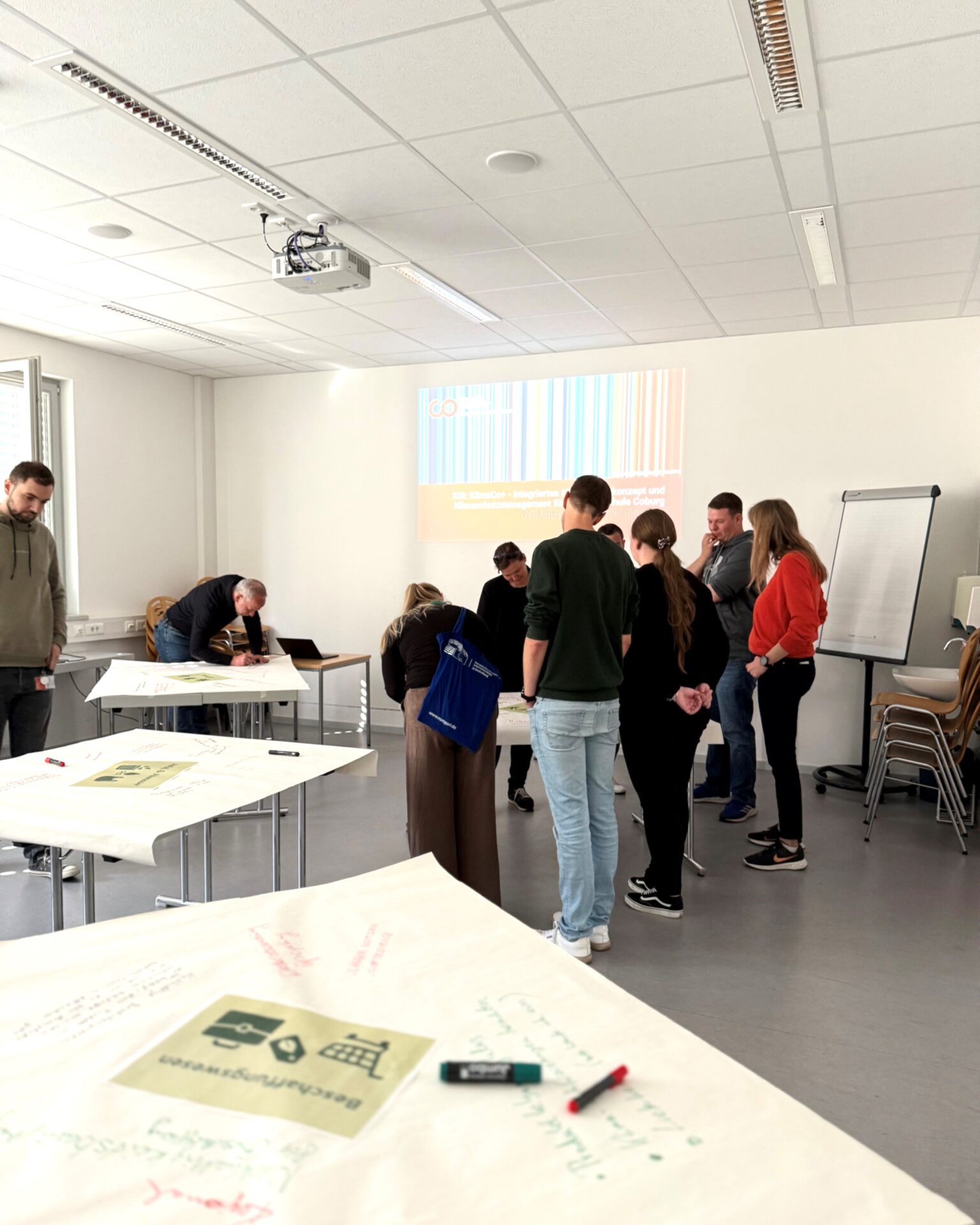 Eine Gruppe von Menschen steht in einem hellen Hörsaal der Hochschule Coburg an einem Tisch und diskutiert. Eine Präsentation zum Thema Klimaneutralität 2040 wird an die Wand projiziert, während Notizen und Diagramme auf Papier sichtbar sind, während die Teilnehmer schreiben oder lesen. at Hochschule Coburg