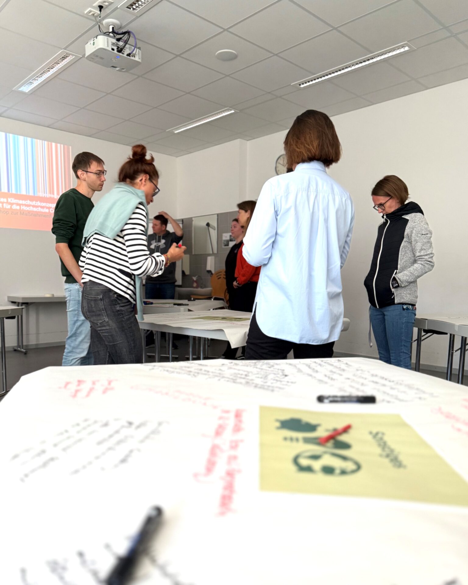 Fünf Personen stehen in einem hellen Hörsaal der Hochschule Coburg an Tischen und unterhalten sich. Zettel mit handschriftlichen Notizen und einem gedruckten Design sind zu sehen, während ein Projektor bunte Streifen, Text und das Ziel der Klimaneutralität bis 2040 projiziert. at Hochschule Coburg