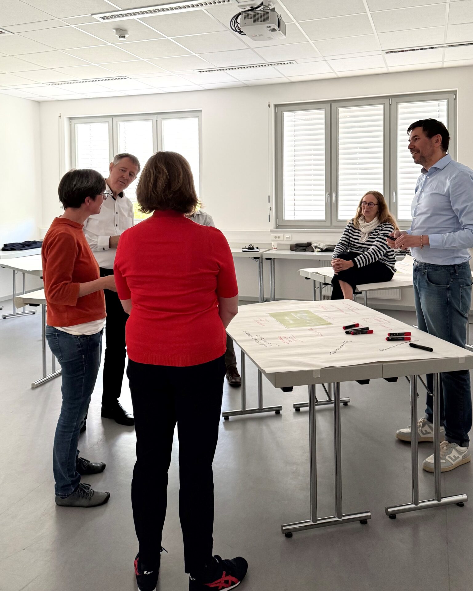 Fünf Erwachsene stehen in einem hellen, modernen Hörsaal der Hochschule Coburg mit Papieren und Stiften an einem Tisch und unterhalten sich. Einige diskutieren über Klimaneutralität 2040, während eine Frau am Tisch sitzt und zuhört. at Hochschule Coburg