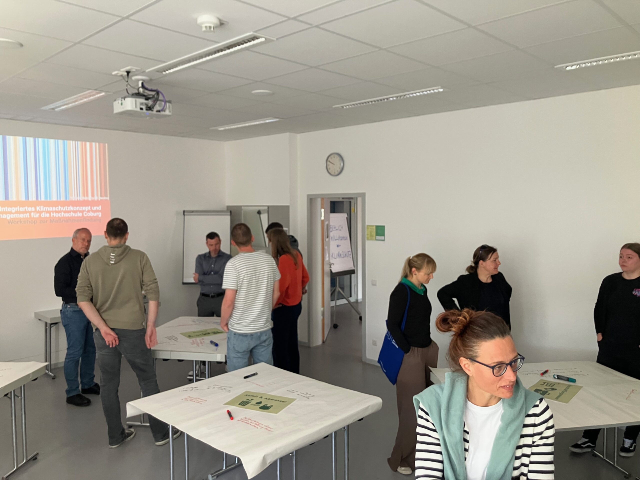 Eine Gruppe von Menschen steht in einem hellen Hörsaal der Hochschule Coburg an Tischen und unterhält sich. Einige lesen in den auf den Tischen liegenden Unterlagen, eine Präsentation zum Thema „Klimaneutralität bis 2040“ wird an die Wand projiziert, und über einer Tür ist eine Uhr zu sehen. at Hochschule Coburg