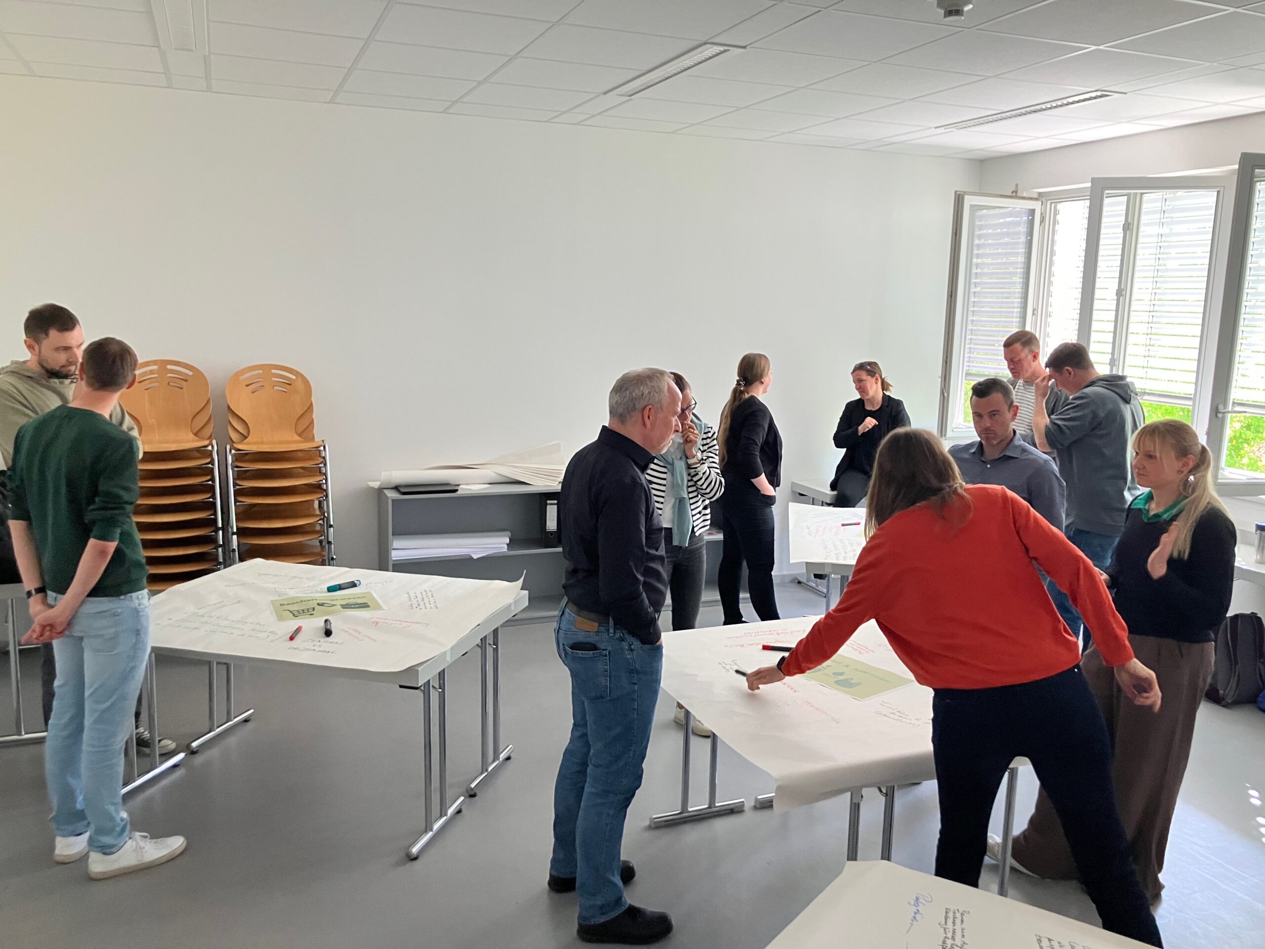 Eine Gruppe von Menschen steht in einem hellen Raum der Hochschule Coburg an mit großen Blättern bedeckten Tischen und unterhält sich. Offenbar arbeiten sie gemeinsam an einem Projekt oder Workshop zum Thema Klimaneutralität und den Zielen für 2040. at Hochschule Coburg