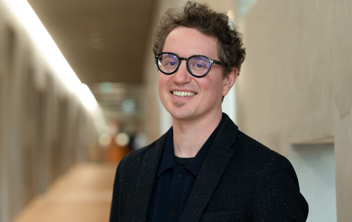 Eine lächelnde Person mit lockigem braunem Haar und Brille, die einen schwarzen Blazer und ein schwarzes Hemd trägt, steht in einem hell erleuchteten Flur mit hellen Wänden. at Hochschule Coburg