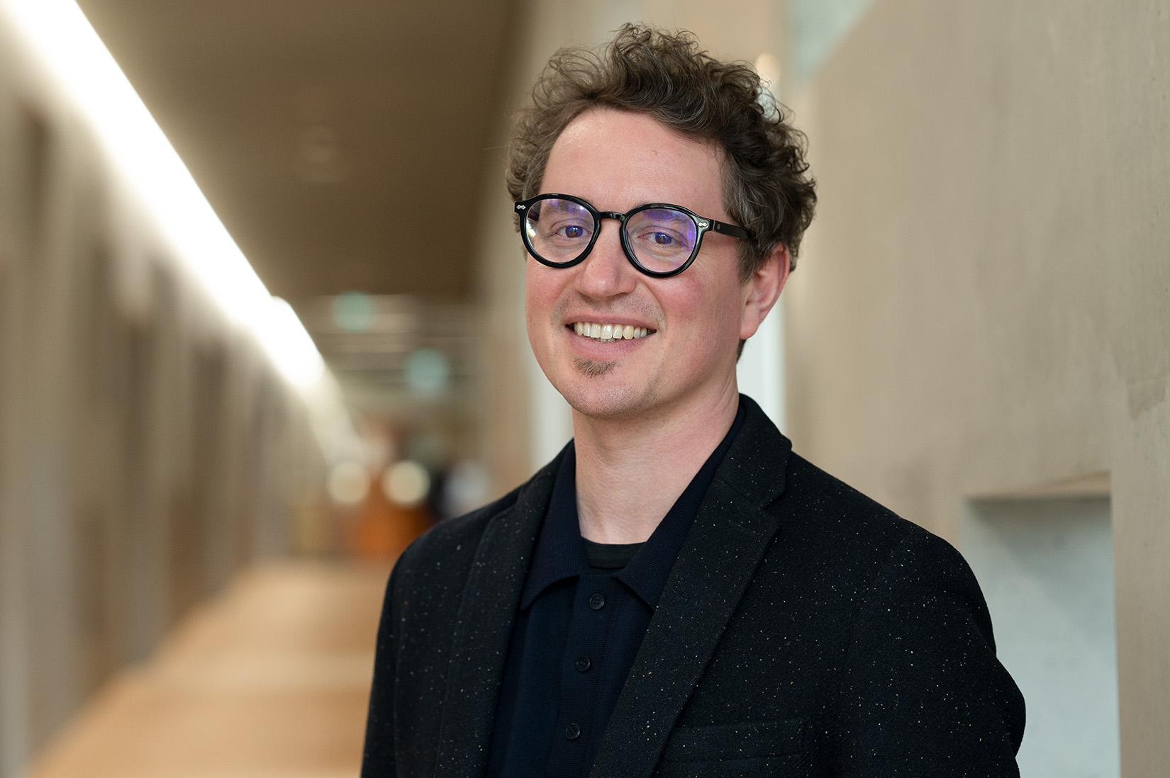 Eine lächelnde Person mit lockigem braunem Haar und Brille, die einen schwarzen Blazer und ein schwarzes Hemd trägt, steht in einem hell erleuchteten Flur mit hellen Wänden. at Hochschule Coburg