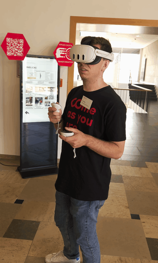 Eine Person mit VR-Headset und Controllern steht in einem Raum auf gefliestem Boden. Sie trägt ein schwarzes T-Shirt mit rotem Text. Im Hintergrund sind QR-Code-Schilder und eine Informationsanzeige zu sehen. at Hochschule Coburg