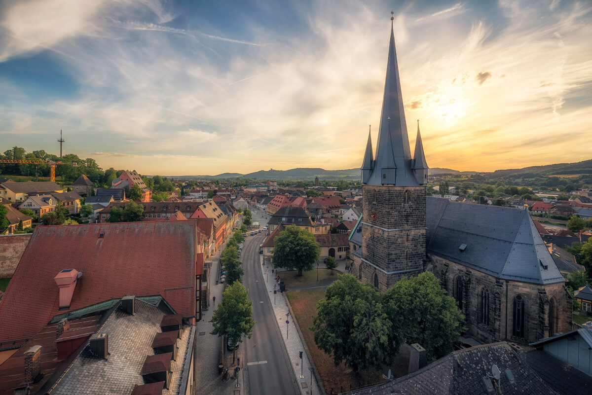 Studieren in Lichtenfels_Stadt Lichtenfels_Foto Klaus Wendel Aerial view of a European town at sunset, featuring a large stone church with a tall spire, surrounding buildings with red-tiled roofs, a winding street, and distant hills under a partly cloudy sky. at Hochschule Coburg