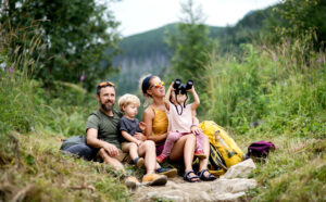 Eine vierköpfige Familie sitzt auf einem Wanderweg im Grünen. Die Eltern und zwei kleine Kinder ruhen auf Felsen; ein Kind schaut durch ein Fernglas. Neben ihnen steht ein gelber Rucksack, im Hintergrund sind Berge zu sehen. at Hochschule Coburg