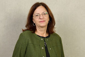 Eine Frau mit schulterlangem braunem Haar, Brille, grüner Jacke und schwarzem Oberteil steht vor einer schlichten hellen Wand und blickt mit neutralem Gesichtsausdruck in die Kamera, was auf eine organisierte und professionelle Präsenz hindeutet. at Hochschule Coburg