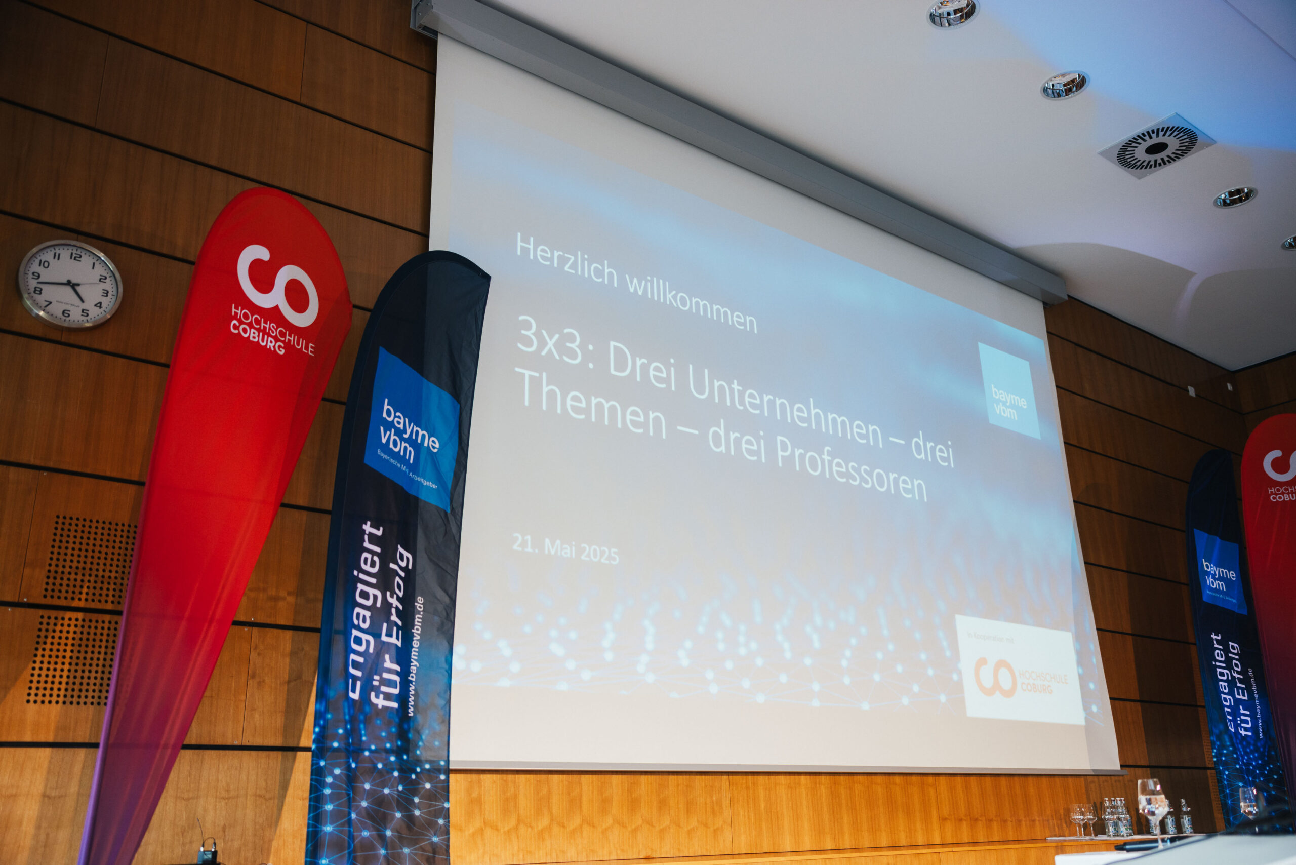 Eine Präsentationsleinwand zeigt den Titel „3x3: Drei Unternehmen – drei Themen – drei Professoren“ mit dem Datum 21. Mai 2025 in einem modernen Hörsaal und hebt den Austausch zwischen Wirtschaft und Wissenschaft neben roten und blauen Event-Bannern hervor. at Hochschule Coburg