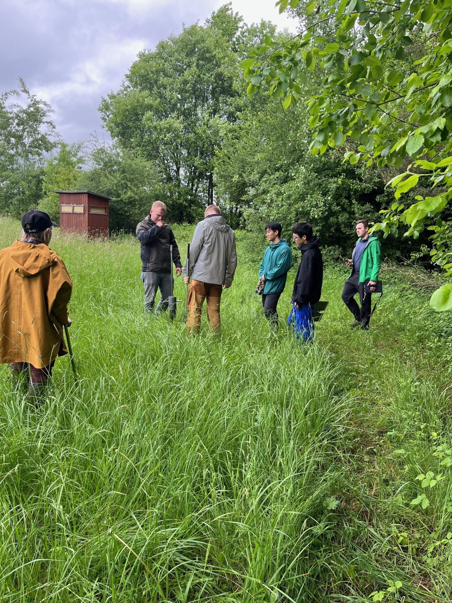 Eine Gruppe von Menschen steht auf einem grasbewachsenen Flur, umgeben von hohen grünen Bäumen. Einige tragen Regenjacken und halten Regenschirme in der Hand. Der Himmel ist bewölkt, was auf die Forschung für klimarobuste Kommunen nahe einer kleinen Holzkonstruktion im Hintergrund hindeutet. at Hochschule Coburg