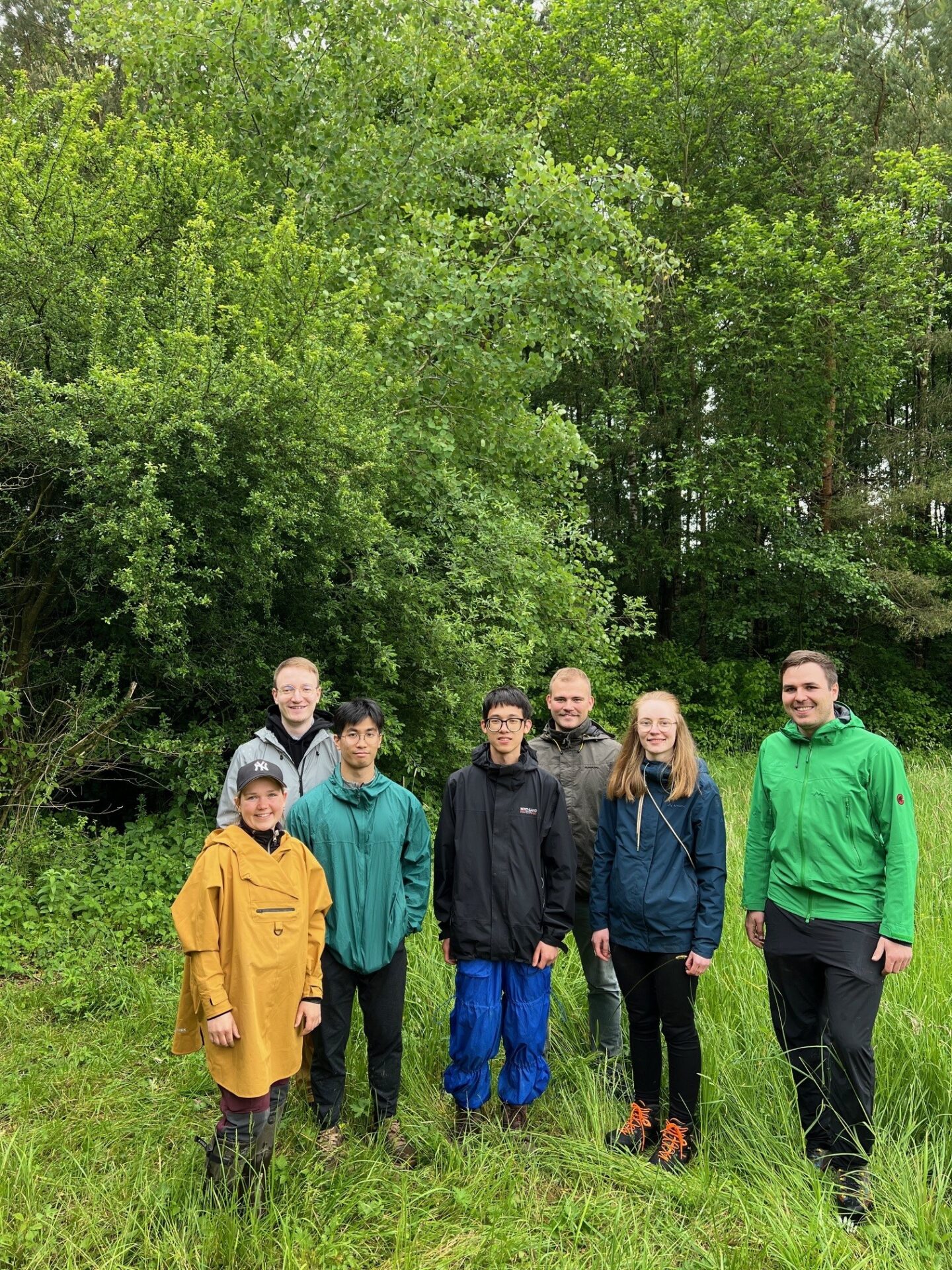 Sieben Menschen stehen in Regenjacken und Outdoor-Kleidung auf einer Wiese vor üppigen grünen Bäumen und lächeln in die Kamera. Ihr Gruppenausflug konzentriert sich auf die Erforschung klimarobuster Kommunen in einem natürlichen, bewaldeten Flur. at Hochschule Coburg