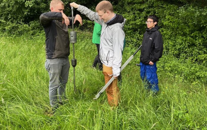 Vier Personen stehen im hohen Gras neben dichten grünen Bäumen. Zwei von ihnen forschen, bedienen ein Bodenprobenentnahmegerät, eine weitere hält eine Schaufel und eine vierte beobachtet in Regenkleidung – eine Feldarbeit im Freien zur Unterstützung klimarobuster Kommunen. at Hochschule Coburg