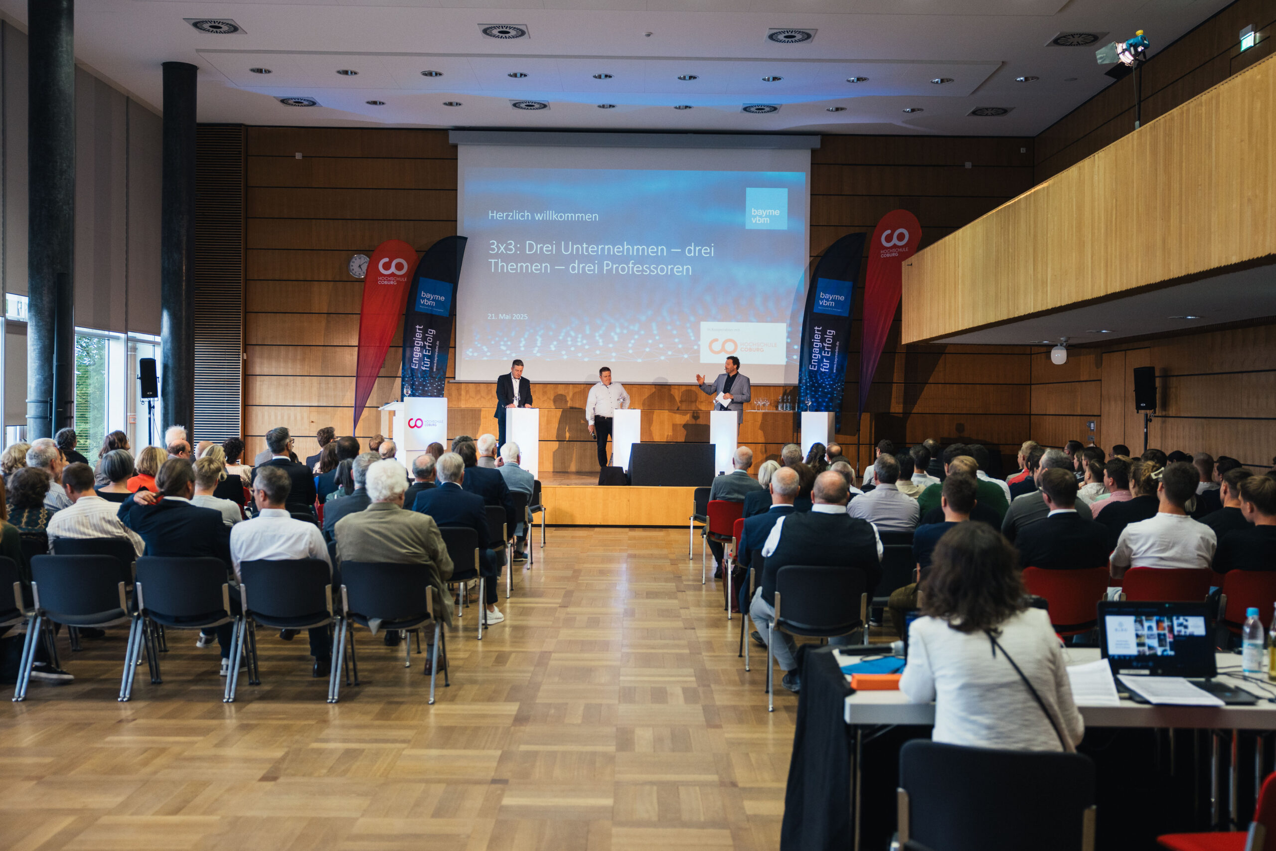 Ein Konferenzraum mit einem Publikum vor einer Bühne, auf der drei Personen sprechen. Auf einem großen Bildschirm dahinter wird eine Begrüßungsbotschaft auf Deutsch mit dem Titel „Einblicke in Wissenschaft“ angezeigt, während an beiden Seiten der Bühne Banner aufgestellt sind. at Hochschule Coburg