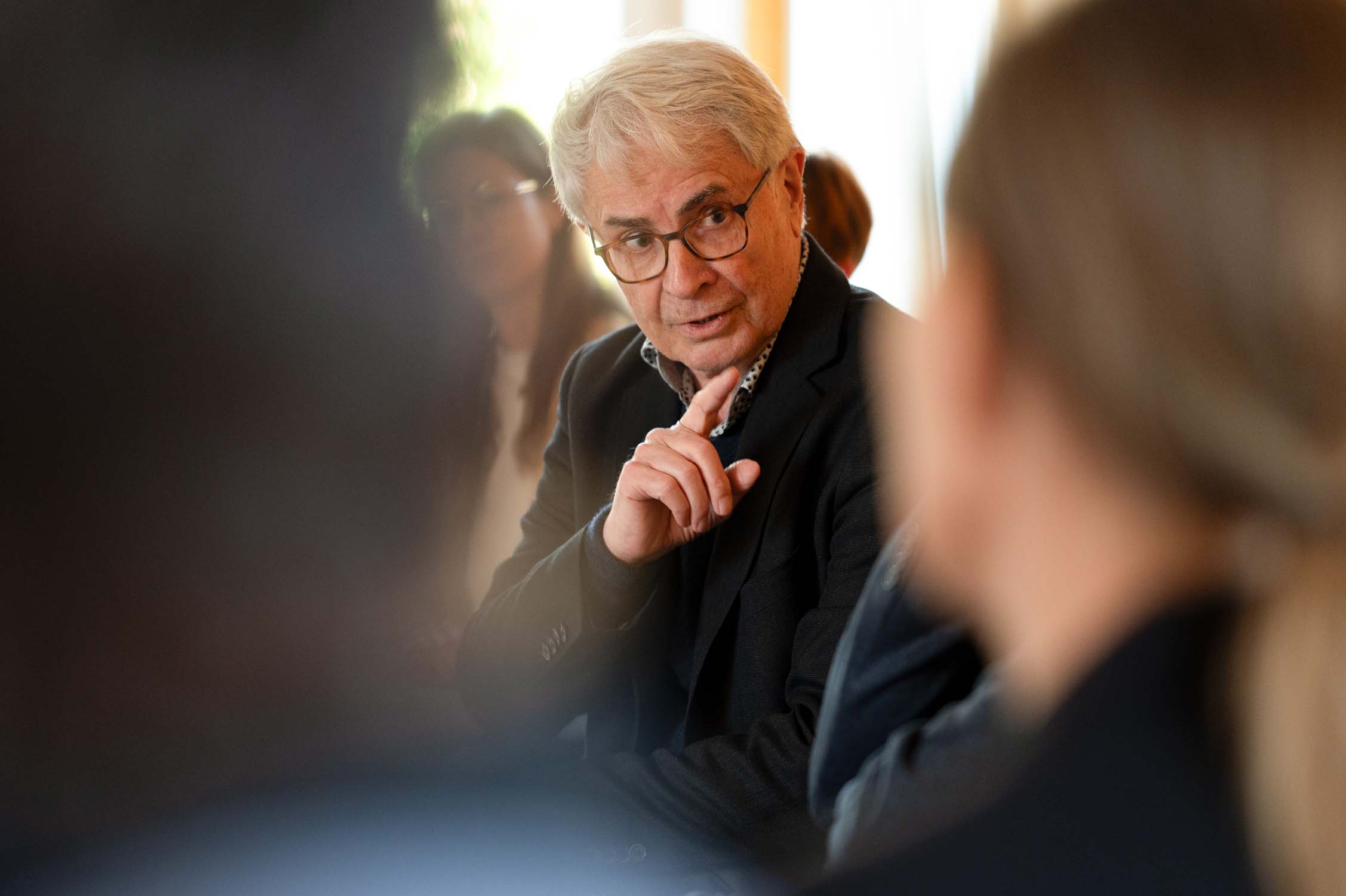 Ein älterer Mann mit grauem Haar und Brille spricht und gestikuliert mit erhobenem Finger, während andere vor einem unscharfen Hintergrund um ihn herum sitzen und eine Gruppendiskussion zum Thema Wissenschaft oder Soziale Arbeit im Rahmen eines IPSG-Treffens vorschlagen. at Hochschule Coburg