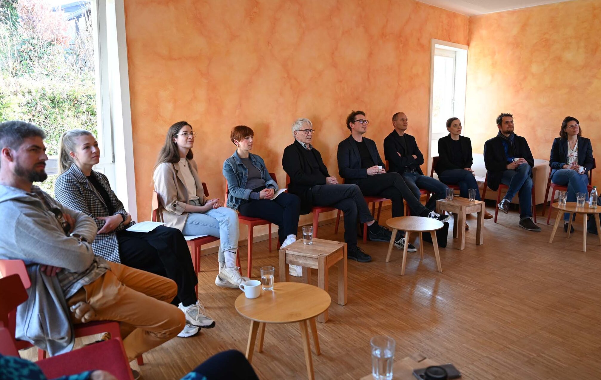 Eine Gruppe von zehn Personen sitzt im Halbkreis auf roten Stühlen in einem hellen Raum mit orangefarbenen Wänden und Holzboden und hört aufmerksam zu – einer spannenden IPSG-Diskussion zum Thema Soziale Arbeit. Vor ihnen stehen kleine Tische mit Getränken und Notizbüchern. at Hochschule Coburg