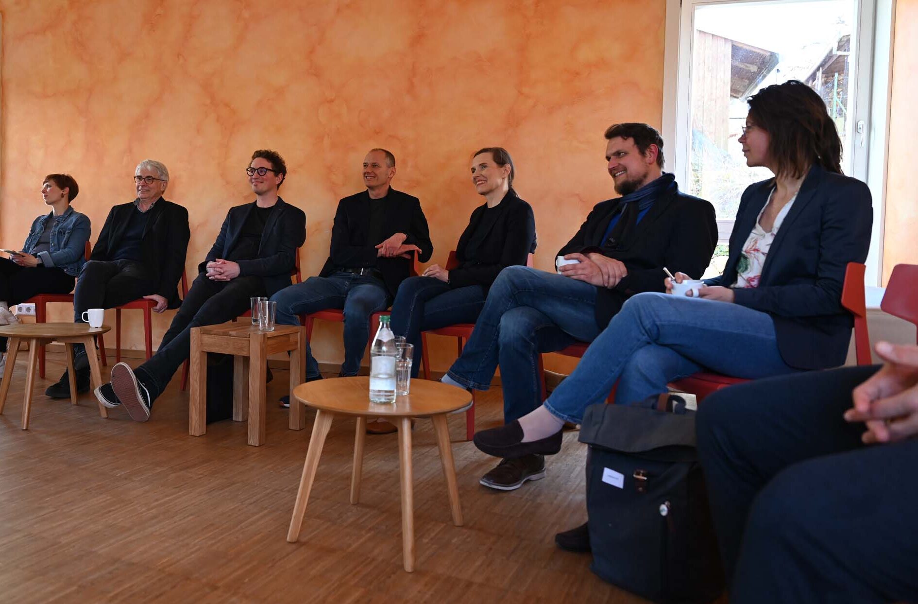 Eine Gruppe von acht Personen sitzt im Halbkreis auf roten Stühlen in einem hellen Raum mit orangefarbenen Wänden und diskutiert mit der IPSG über Soziale Arbeit. Vor ihnen stehen kleine Holztische mit Wasserflaschen und Gläsern. at Hochschule Coburg