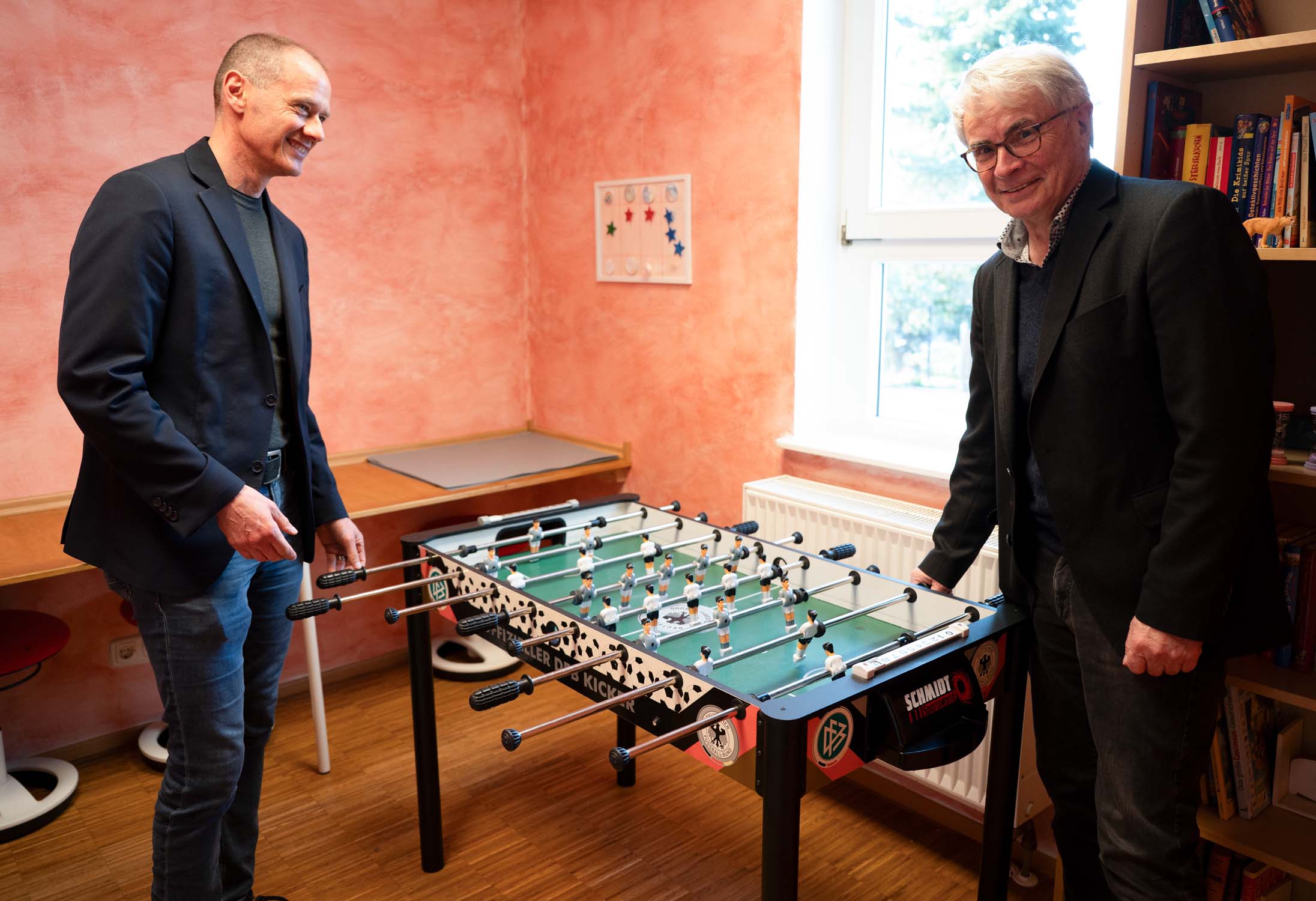 Zwei Männer stehen und lächeln, während sie Tischfußball in einem gemütlichen Raum mit pfirsichfarbenen Wänden, einem Fenster, einem Bücherregal und einem kleinen Schreibtisch spielen – ein Spiegelbild der freundlichen Atmosphäre, die oft in Einrichtungen der sozialen Arbeit oder Wissenschaft wie der IPSG herrscht. at Hochschule Coburg