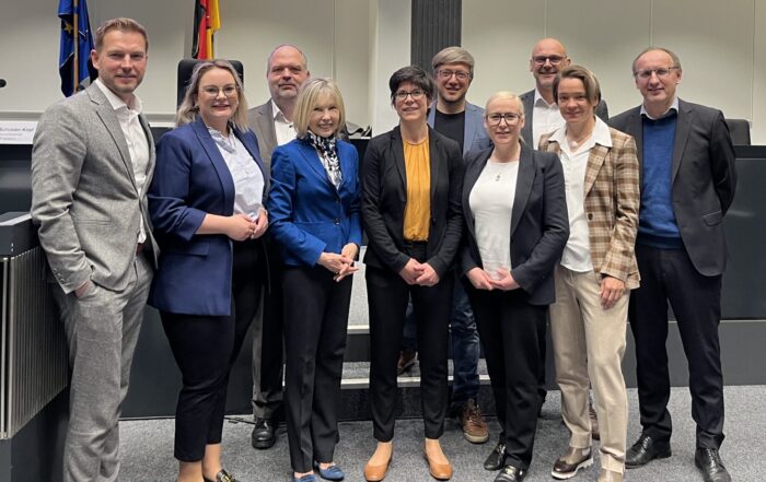 Eine Gruppe von elf Personen steht lächelnd in einem modernen Konferenzraum mit schwarz-weißem Dekor zusammen, der die Themen Wirtschaft und Migration beleuchtet. Im Hintergrund sind zwei Flaggen und ein großer Bildschirm mit blauem Display zu sehen. at Hochschule Coburg