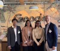 Four people in business attire stand smiling in front of a Google sign on a vintage map wall background, inside an office setting. at Hochschule Coburg