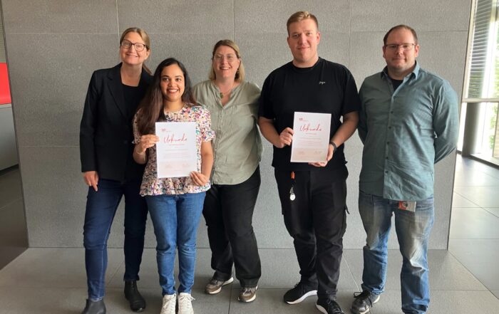 Fünf Personen stehen in einem Innenraum vor einer grauen Wand, zwei in der Mitte halten Urkunden in der Hand und lächeln. Die leger gekleidete Gruppe wirkt fröhlich, was auf Doktorandinnen und Doktoranden schließen lässt, die eine Leistung an der Hochschule Coburg feiern. at Hochschule Coburg