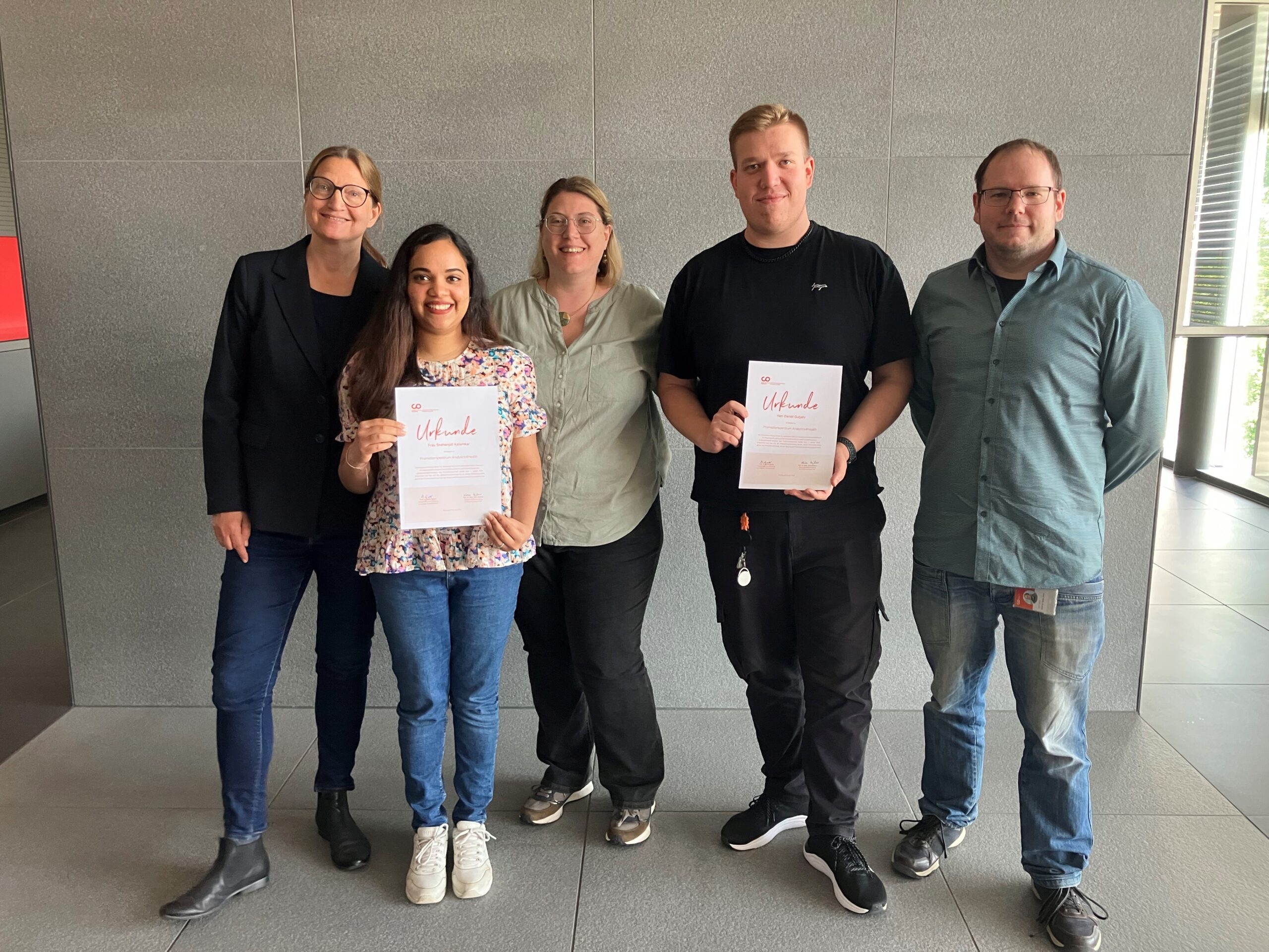 Fünf Personen stehen in einem Innenraum vor einer grauen Wand, zwei in der Mitte halten Urkunden in der Hand und lächeln. Die leger gekleidete Gruppe wirkt fröhlich, was auf Doktorandinnen und Doktoranden schließen lässt, die eine Leistung an der Hochschule Coburg feiern. at Hochschule Coburg