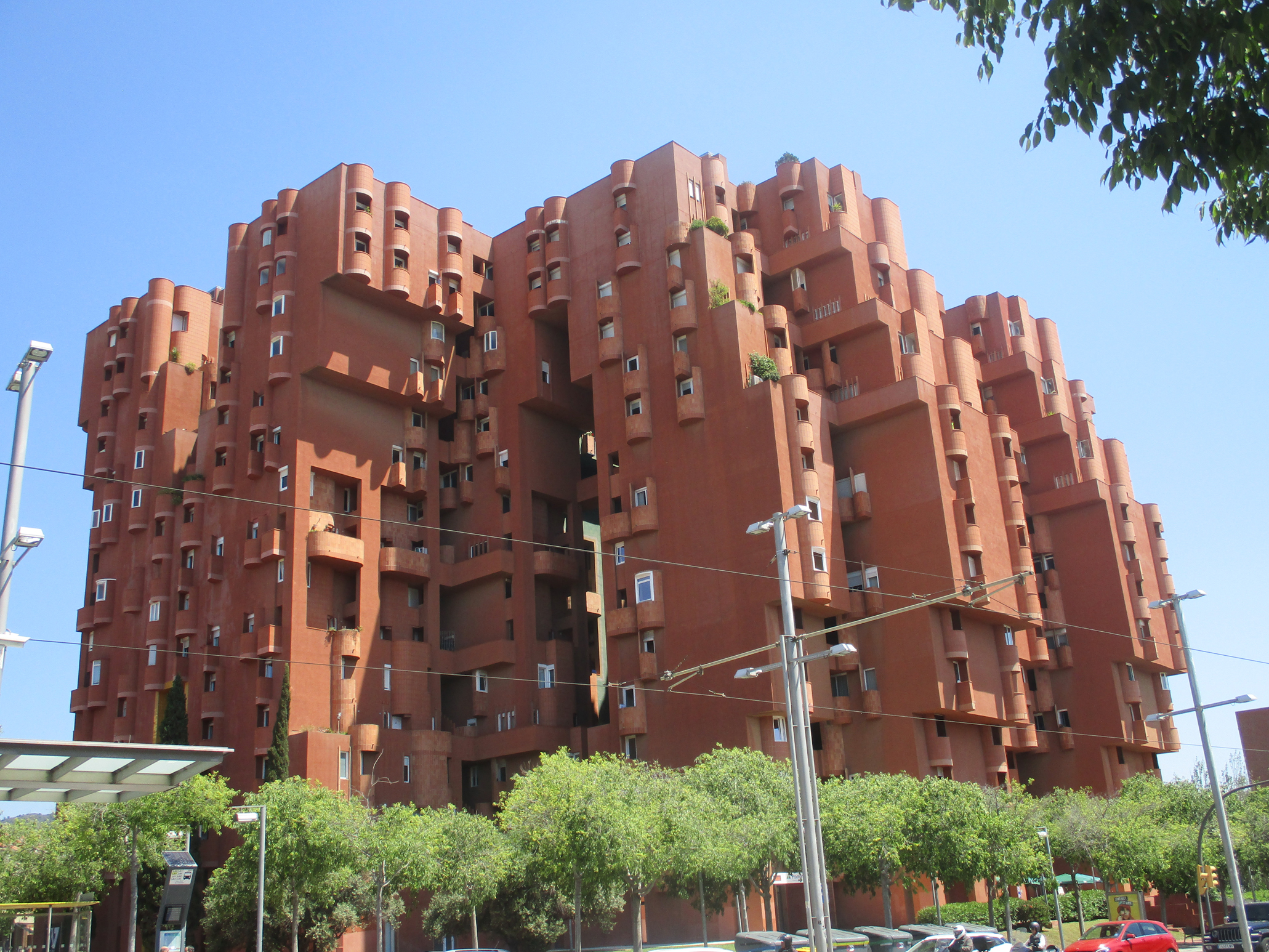Ein großes, rotbraunes, modernes Wohngebäude mit ungewöhnlicher, blockartiger Architektur und vielen Fenstern steht in Barcelona, umgeben von grünen Bäumen und geparkten Autos unter einem klaren blauen Himmel. at Hochschule Coburg
