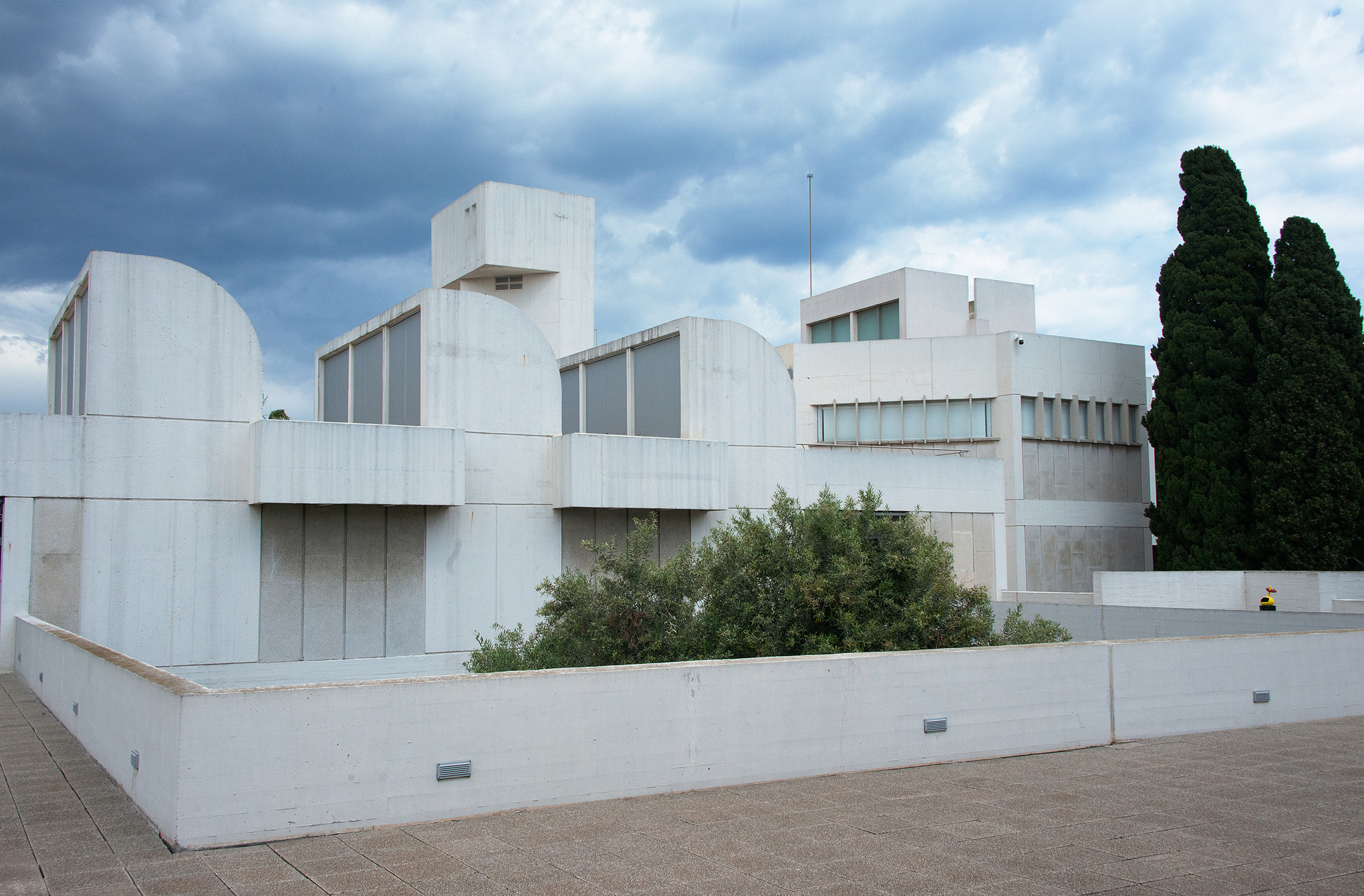 Ein modernes weißes Betongebäude mit geometrischen Formen und flachen Dächern zeigt zeitgenössische Architektur, umgeben von einer niedrigen Mauer und Grün unter einem bewölkten Himmel. Zwei hohe Bäume erheben sich auf der rechten Seite des Gebäudes. at Hochschule Coburg